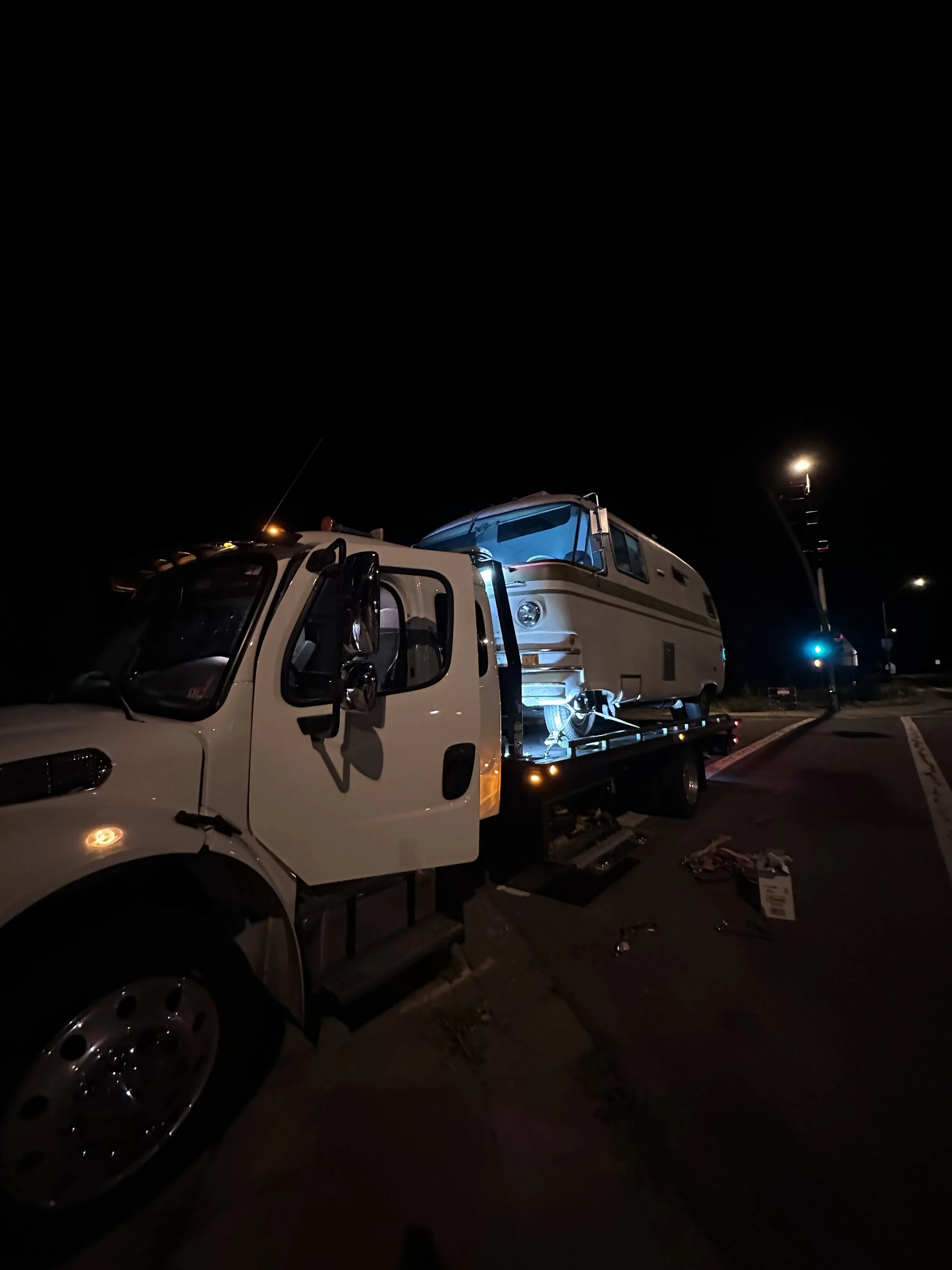 Night scene in a parking lot with a white tow truck carrying a vintage camper trailer, illuminated by streetlights and vehicle lights, with some scattered objects on the ground.