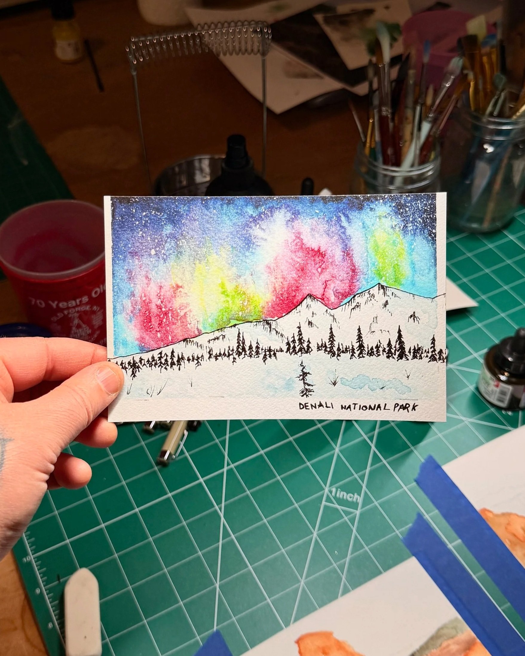 Hand holding a colorful watercolor painting of mountains and trees at Denali National Park, framed by art supplies and workspace tools.