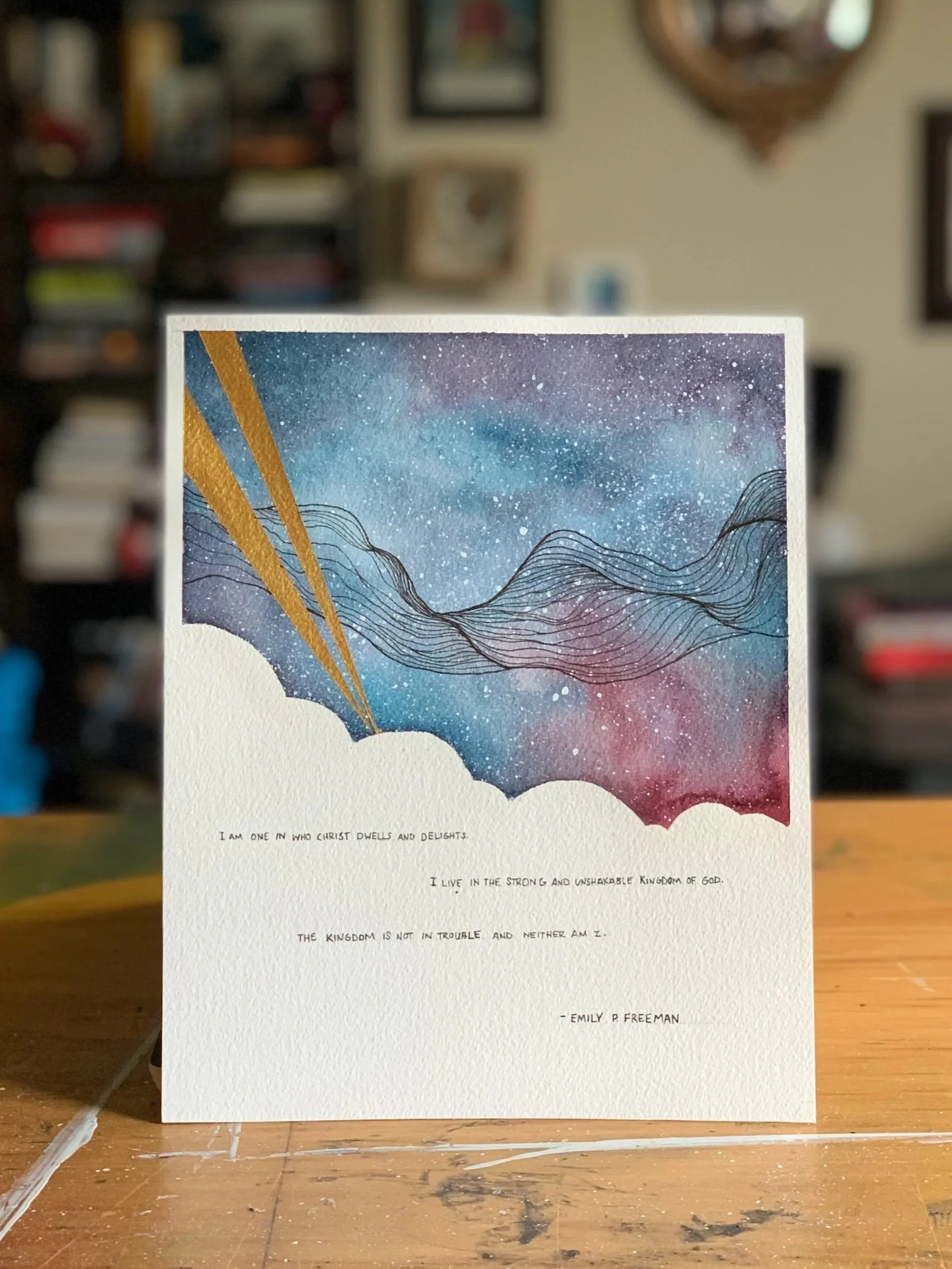 A watercolor painting of a starry night sky with swirling clouds and a mountain range silhouette. Gold diagonal lines cross the sky. Text at the bottom includes a biblical quote by Emily P. Freeman.