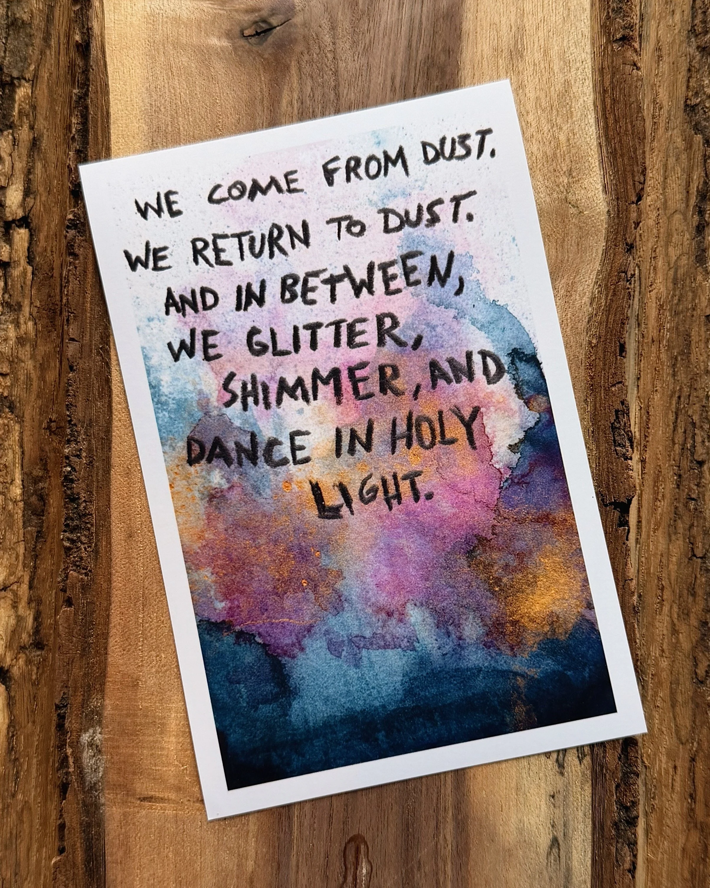 A colorful watercolor painting with a handwritten message that reads: "We come from dust. We return to dust. And in between, we glitter, shimmer, and dance in holy light." The painting features shades of pink, purple, blue, and orange.