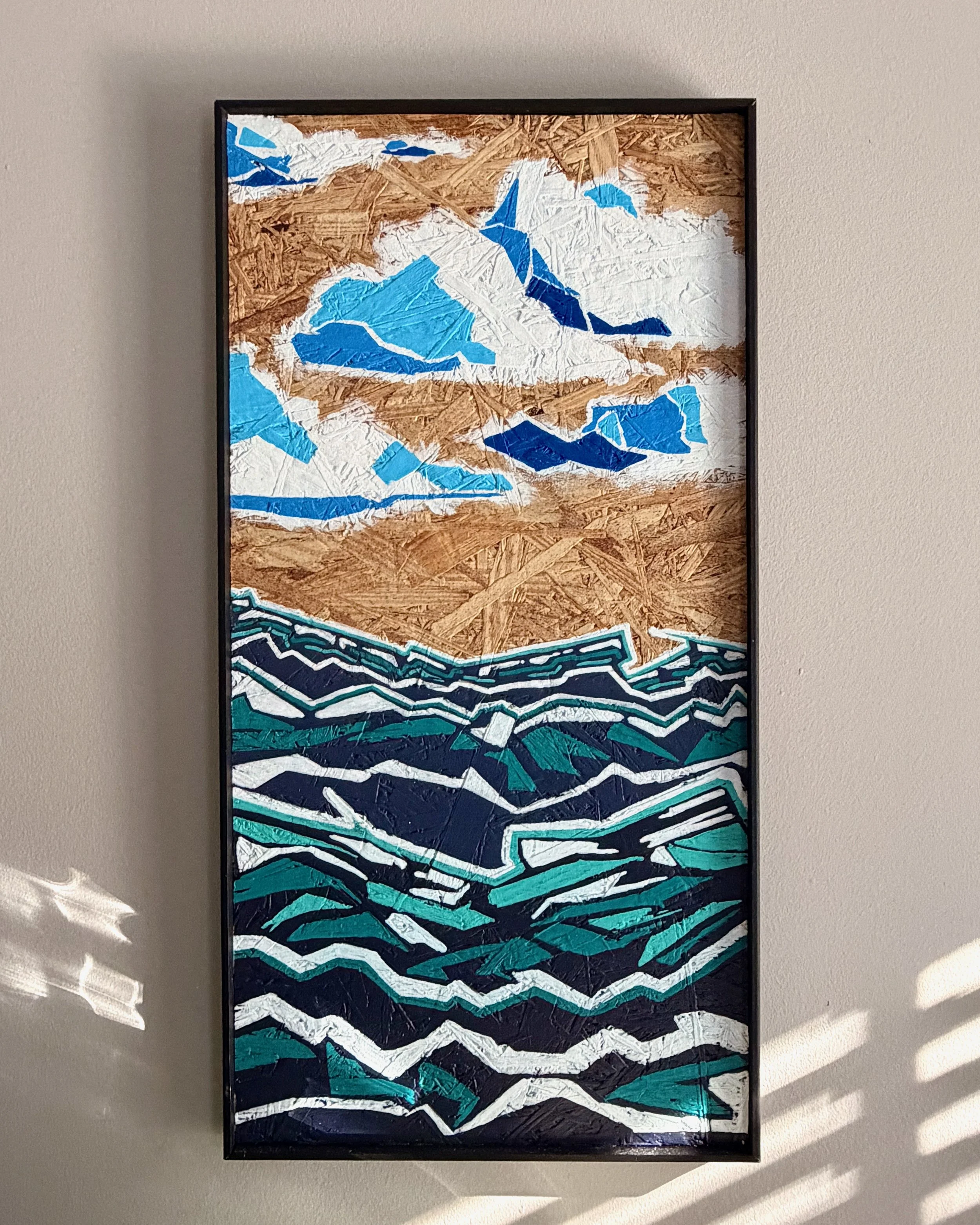 A framed abstract artwork hanging on a wall, featuring jagged, colorful shapes in shades of blue, teal, black, white, brown, and beige, resembling a stylized mountain landscape or water scene.