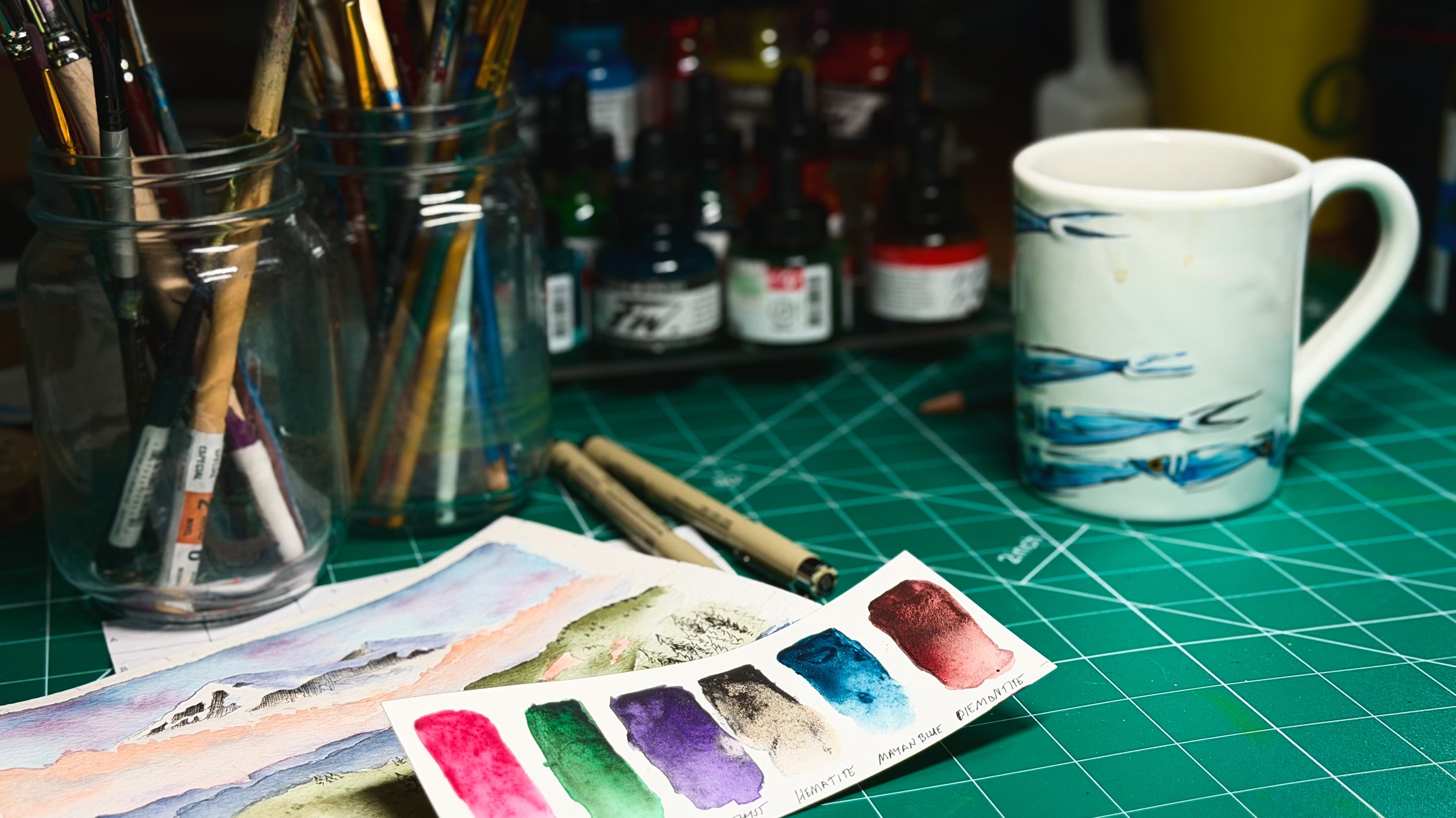 A creative workspace with watercolor swatches, a landscape painting, paintbrushes, ink bottles, pens, and a white mug arranged on a green cutting mat.