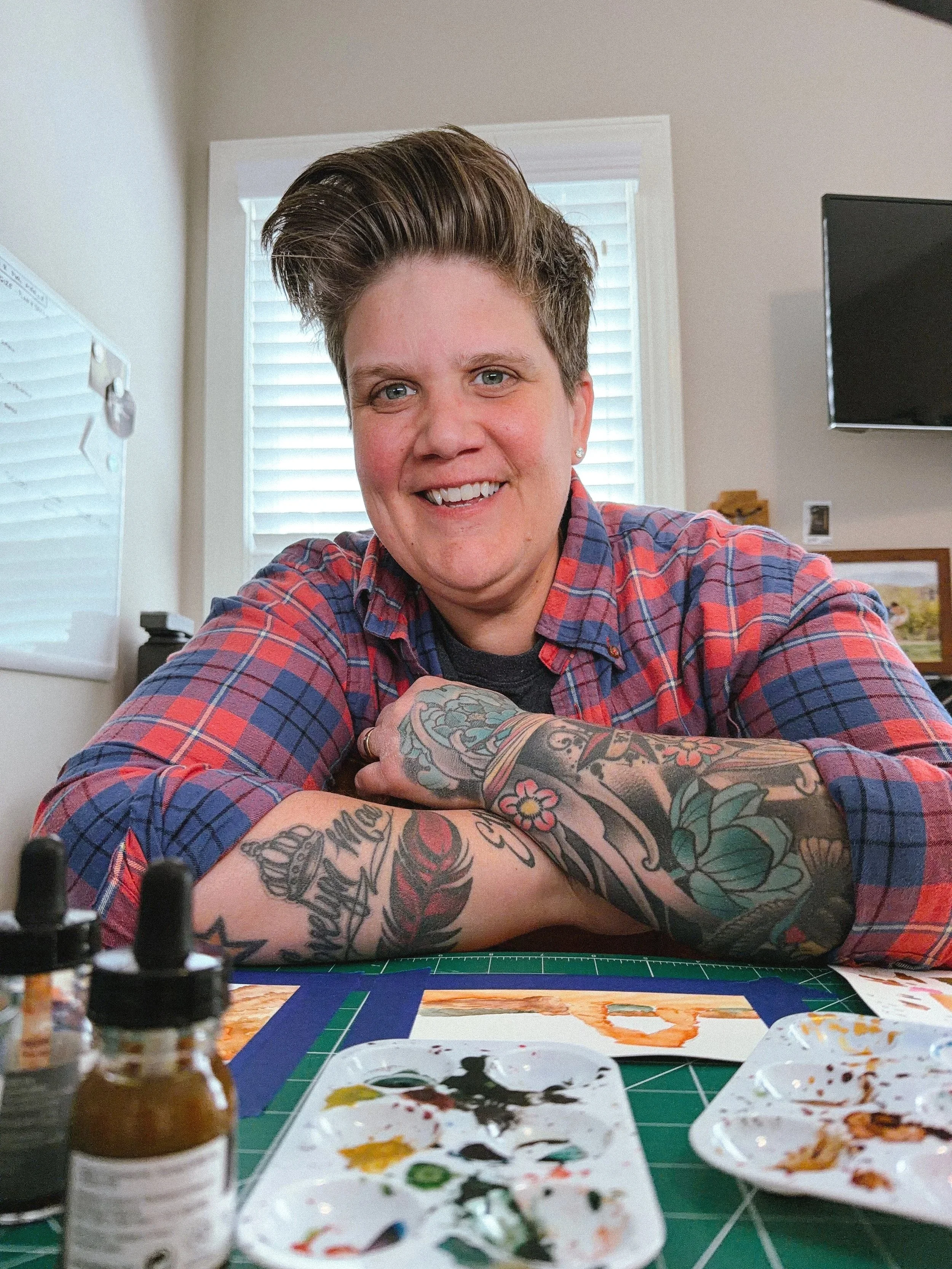 Person smiling at an art desk with tattooed arms crossed, wearing a red and blue plaid shirt. Paint bottles, palettes, watercolor paper, and a green cutting mat are in the foreground, with a bright studio workspace in the background.