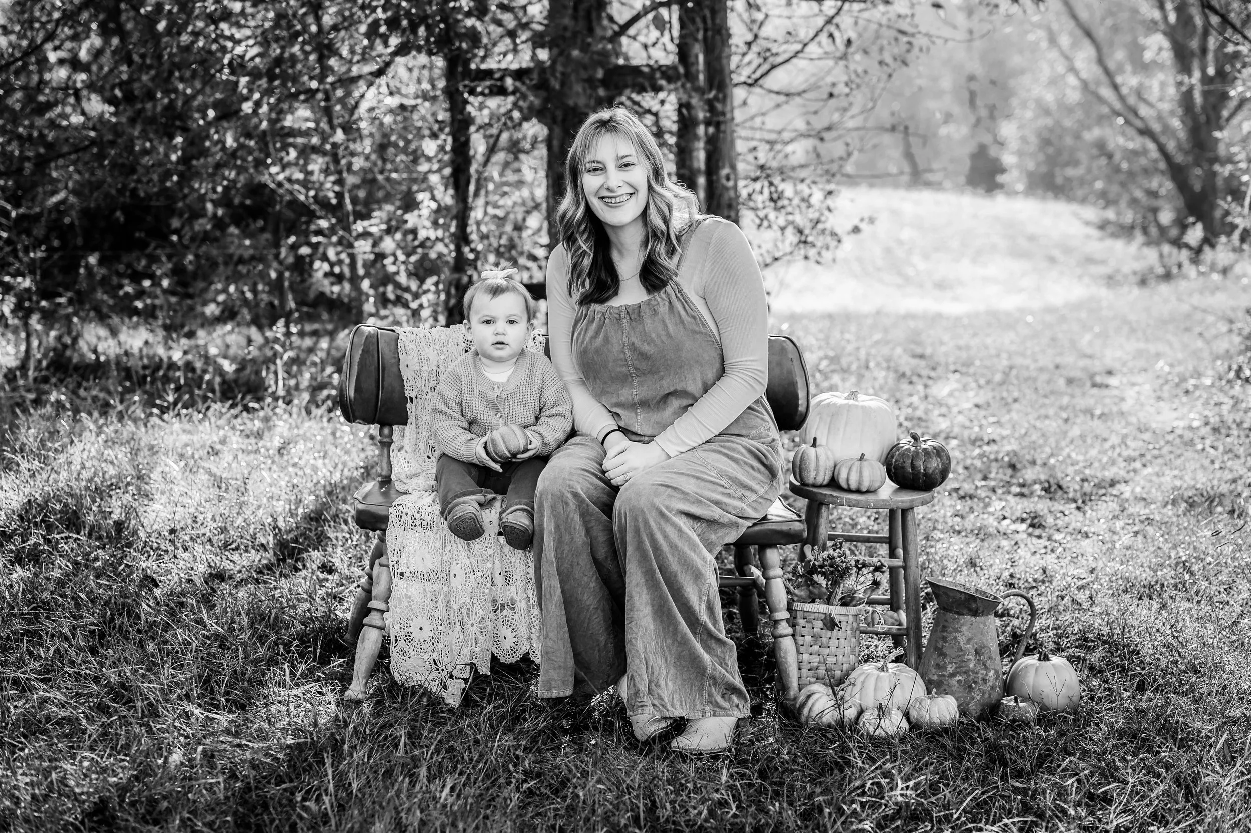 Black and white photo of a woman and a young girl sitting outdoors on a bench, surrounded by pumpkins, with trees and a pathway in the background.