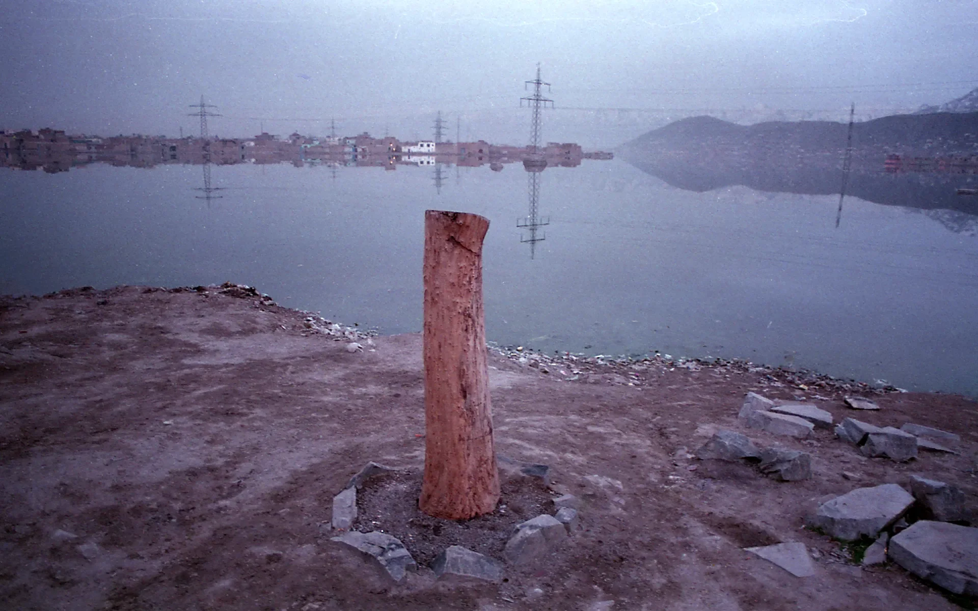 Minimalist documentary landscape featuring a wooden post by a calm lake with mountains in the background, Kabul, Afghanistan.