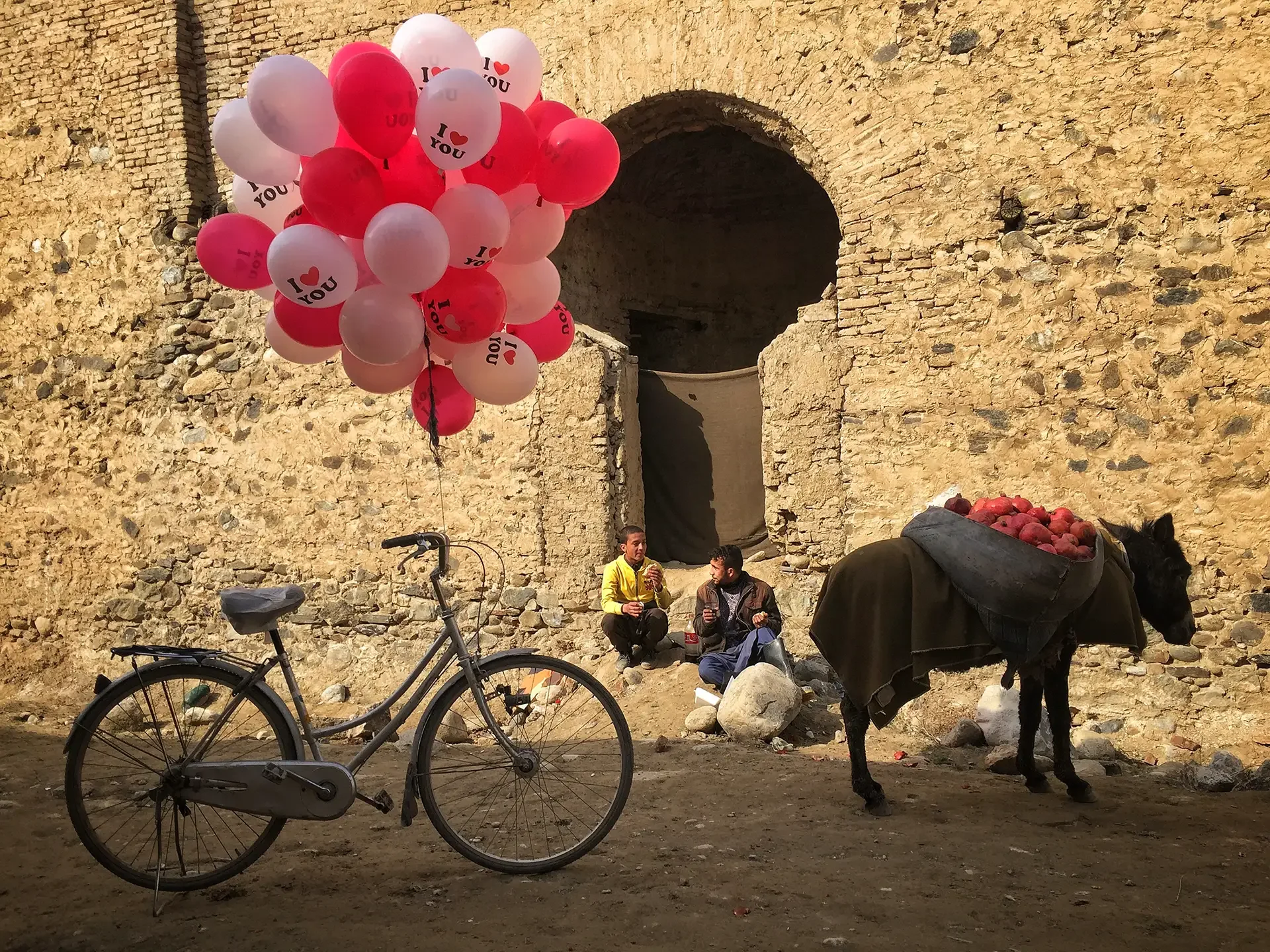 Documentary street photography featuring a bicycle and colorful balloons by an old brick archway,Ghale Fatollah, Kabul,  Afghanistan.