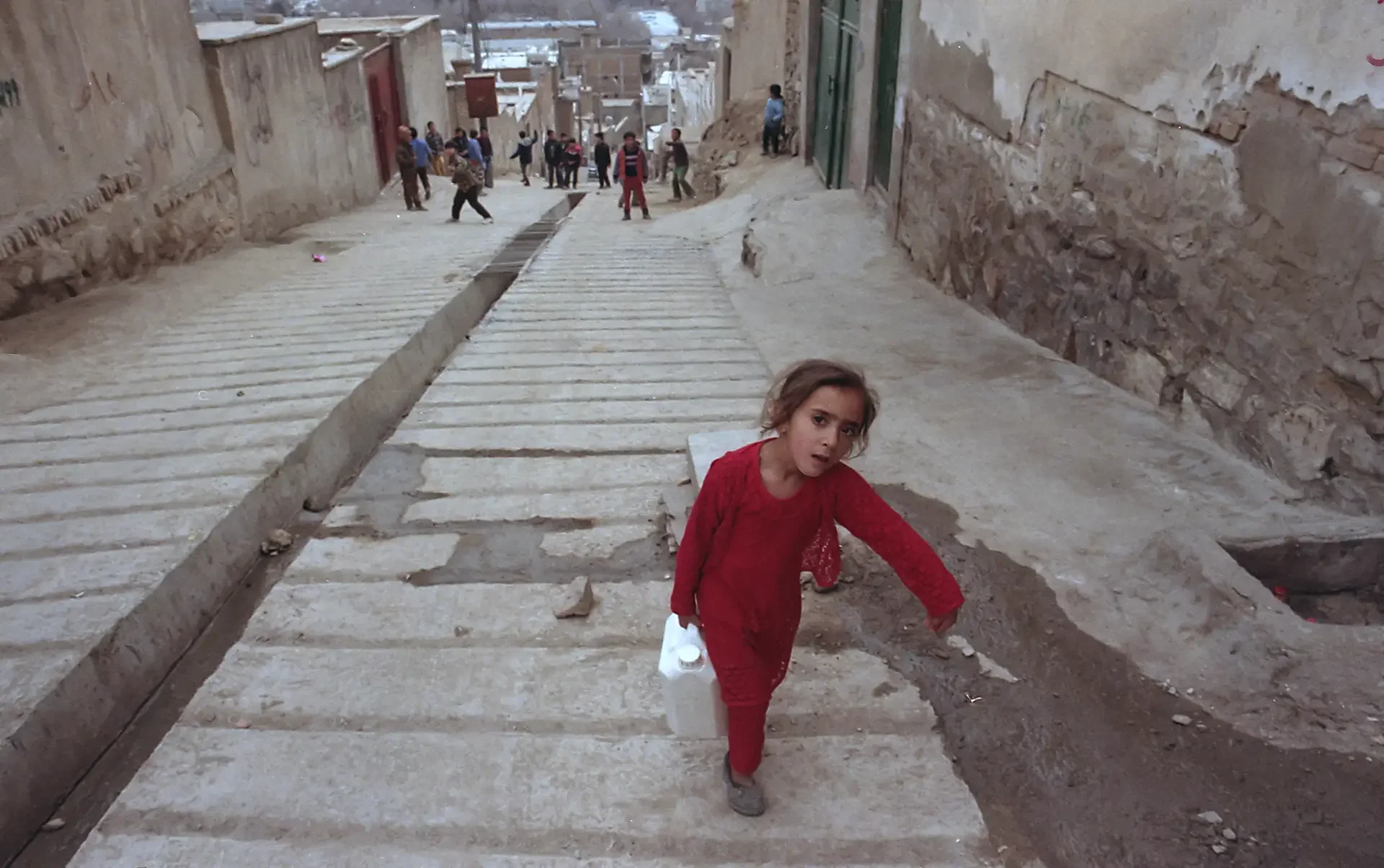 Street photography featuring a little girl walking up a stepped alleyway, carrying a water container home from a long distance, Kabul,  Afghanistan