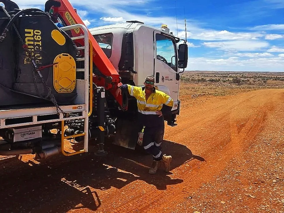 A person in a yellow safety jacket and black pants standing next to a large white utility truck with a crane in a dirt landscape under a partly cloudy sky.