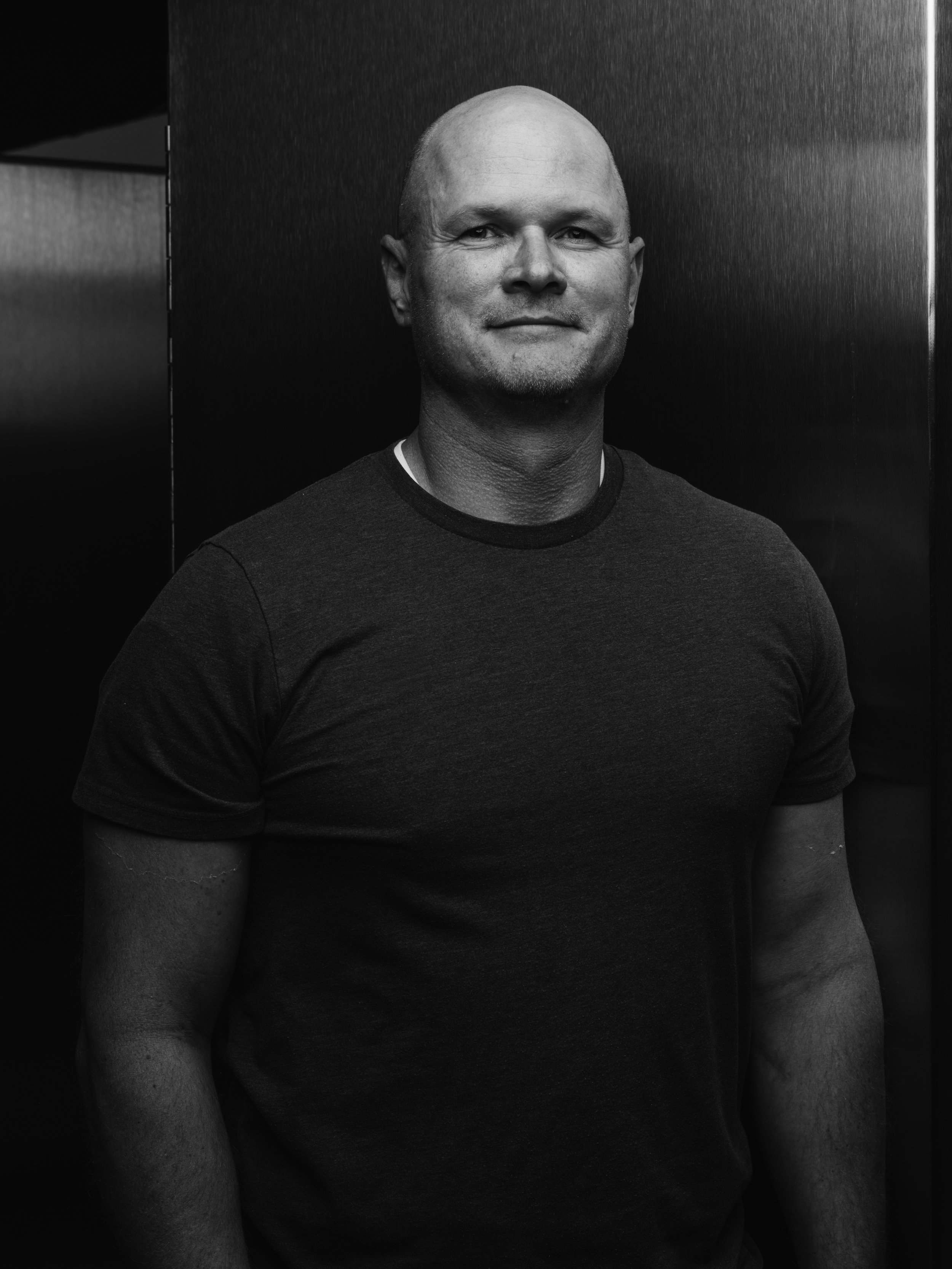 A black and white portrait of a man with a shaved head, smiling slightly, wearing a dark t-shirt, standing against a dark background.