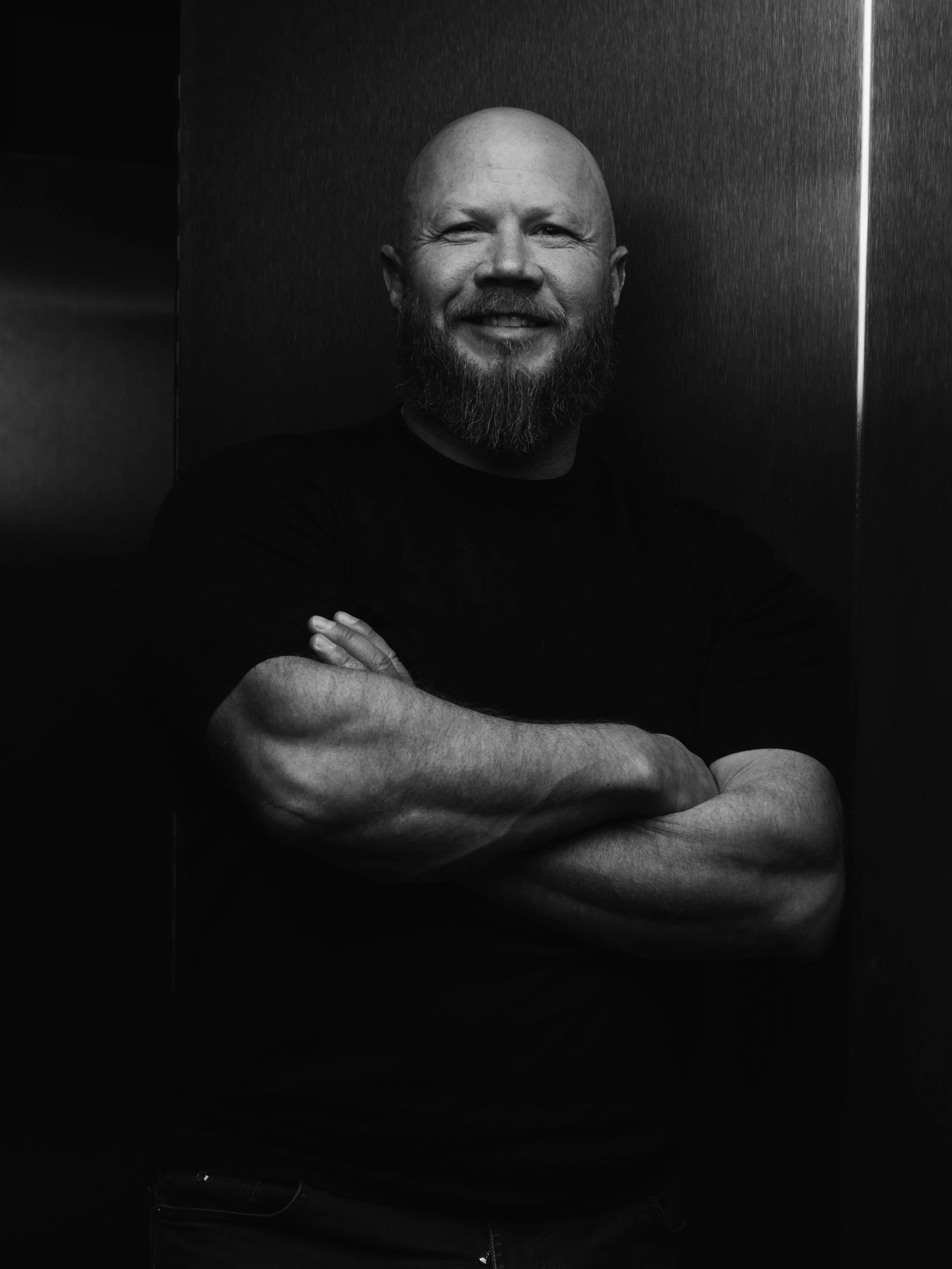 A smiling man with a beard and a shaved head, crossing his arms, standing against a dark textured background.