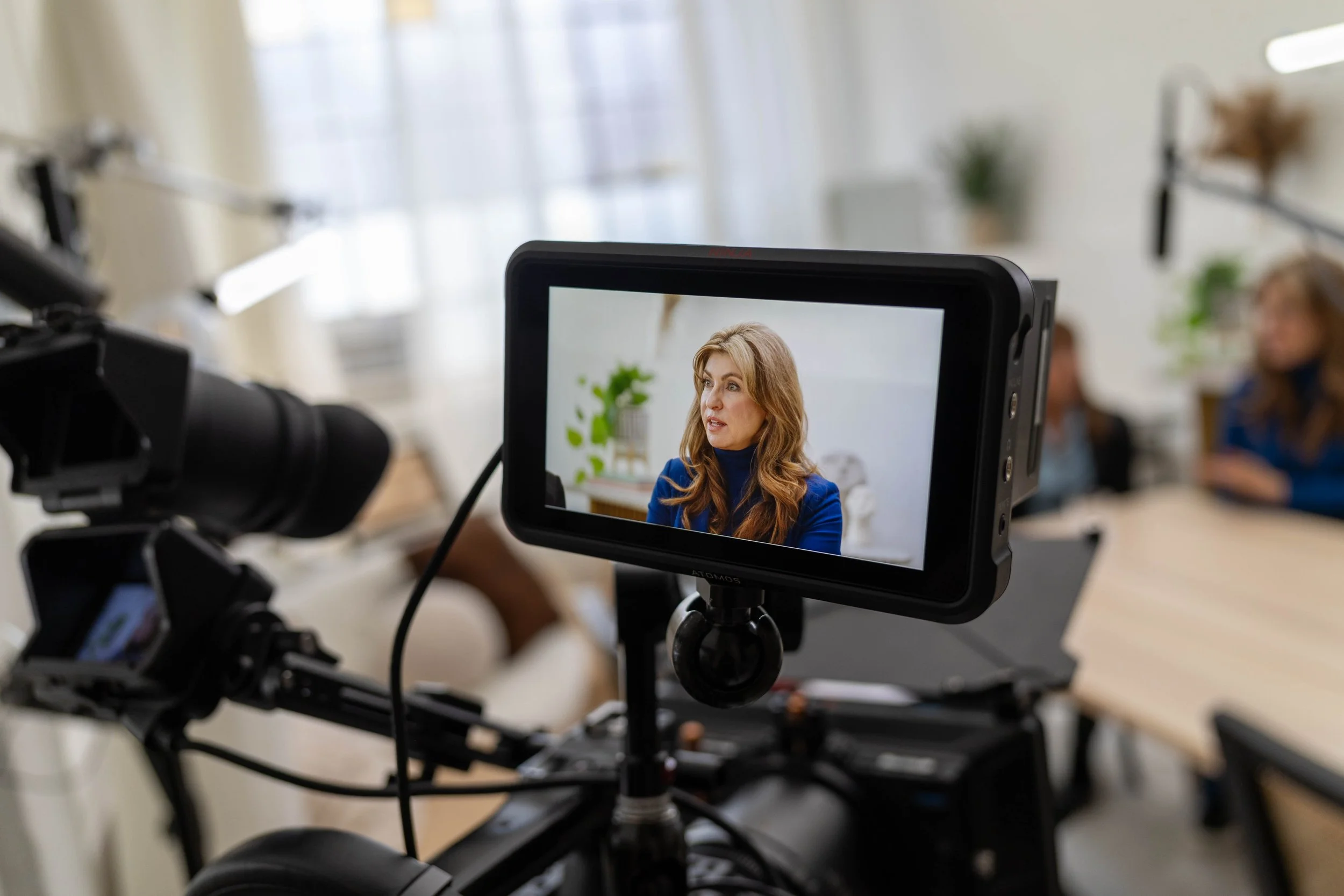 A woman with long wavy hair and a blue jacket is being recorded by a camera with a monitor showing her face. The background is blurred with light coming through windows, and there are other people present.
