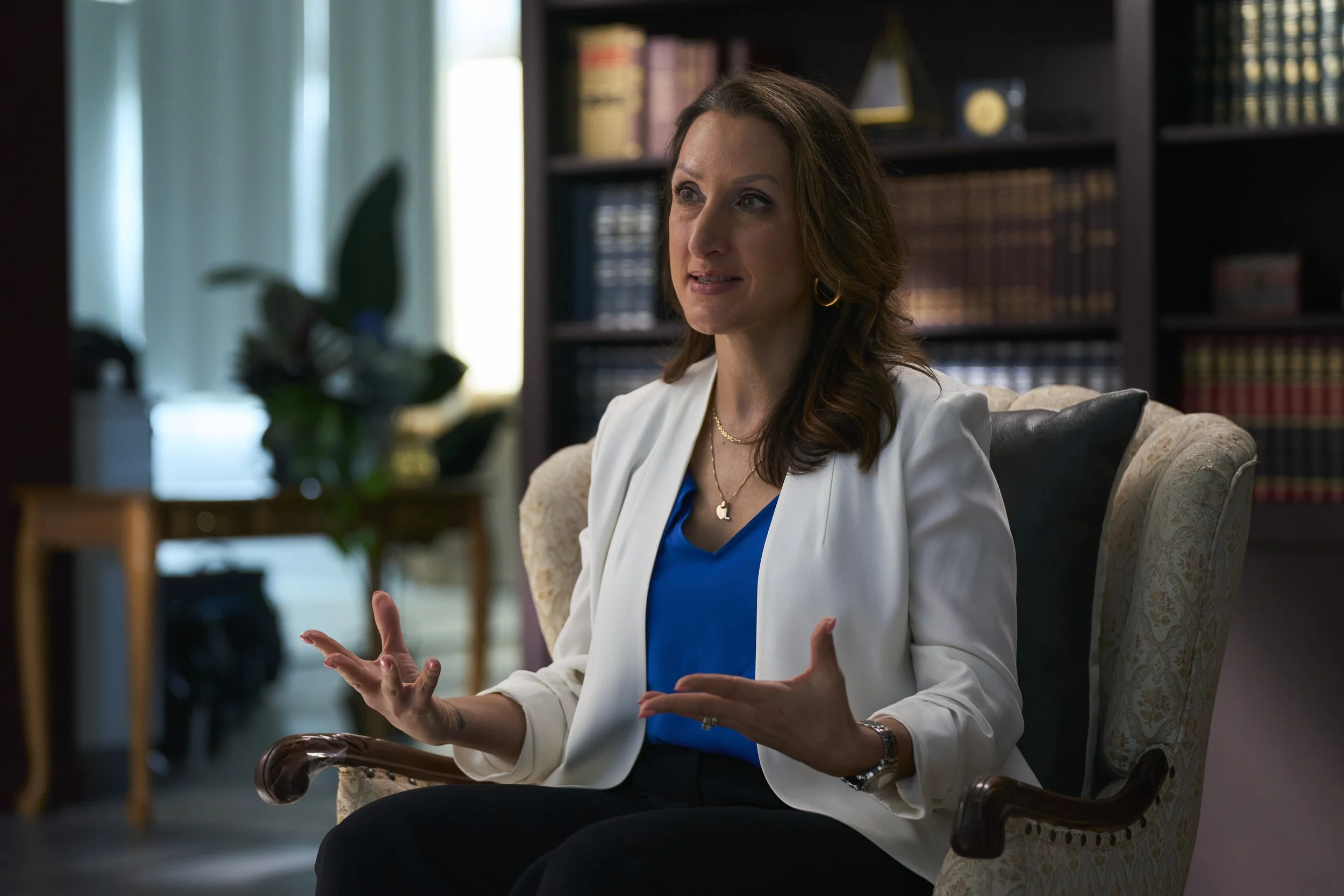 A woman with brown hair wearing a white blazer and blue shirt, sitting and gesturing with her hands in a room with bookcases and a plant in the background.