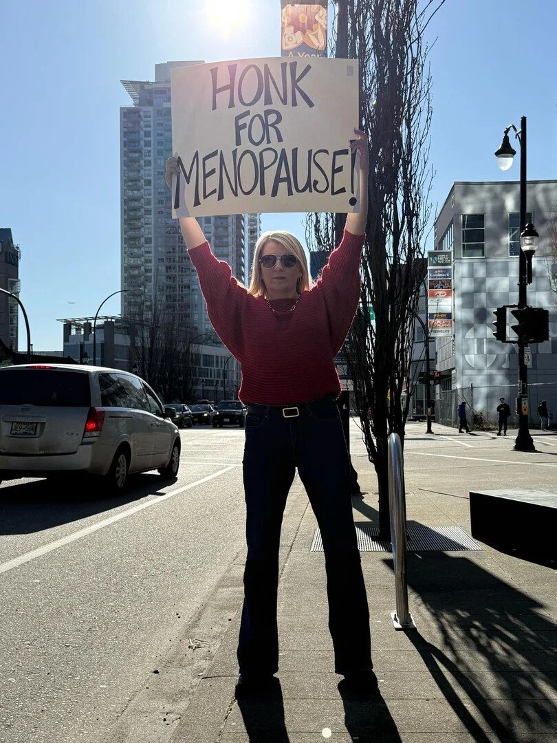 A woman wearing sunglasses, a red sweater, and black pants holds a sign that says "HONK FOR MENOPAUSE!" during daytime in an urban setting with high-rise buildings and cars in the background.