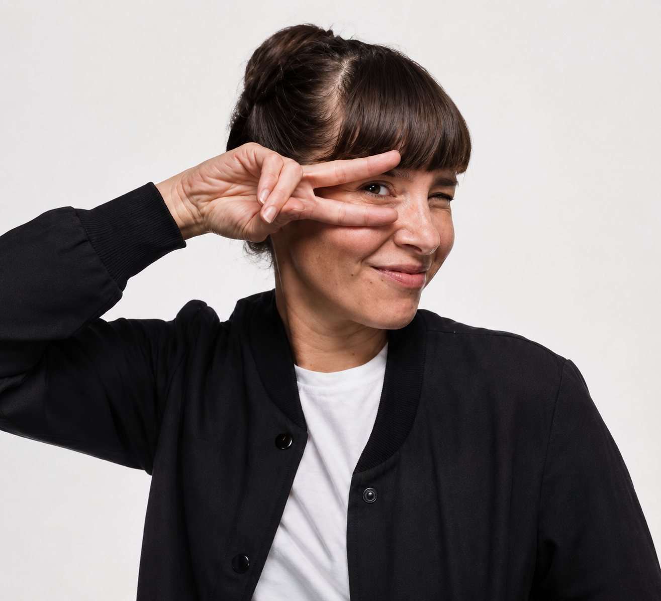 A woman with short brown hair making a peace sign with her right hand near her face, smiling and winking at the camera, wearing a black jacket over a white t-shirt against a plain white background.
