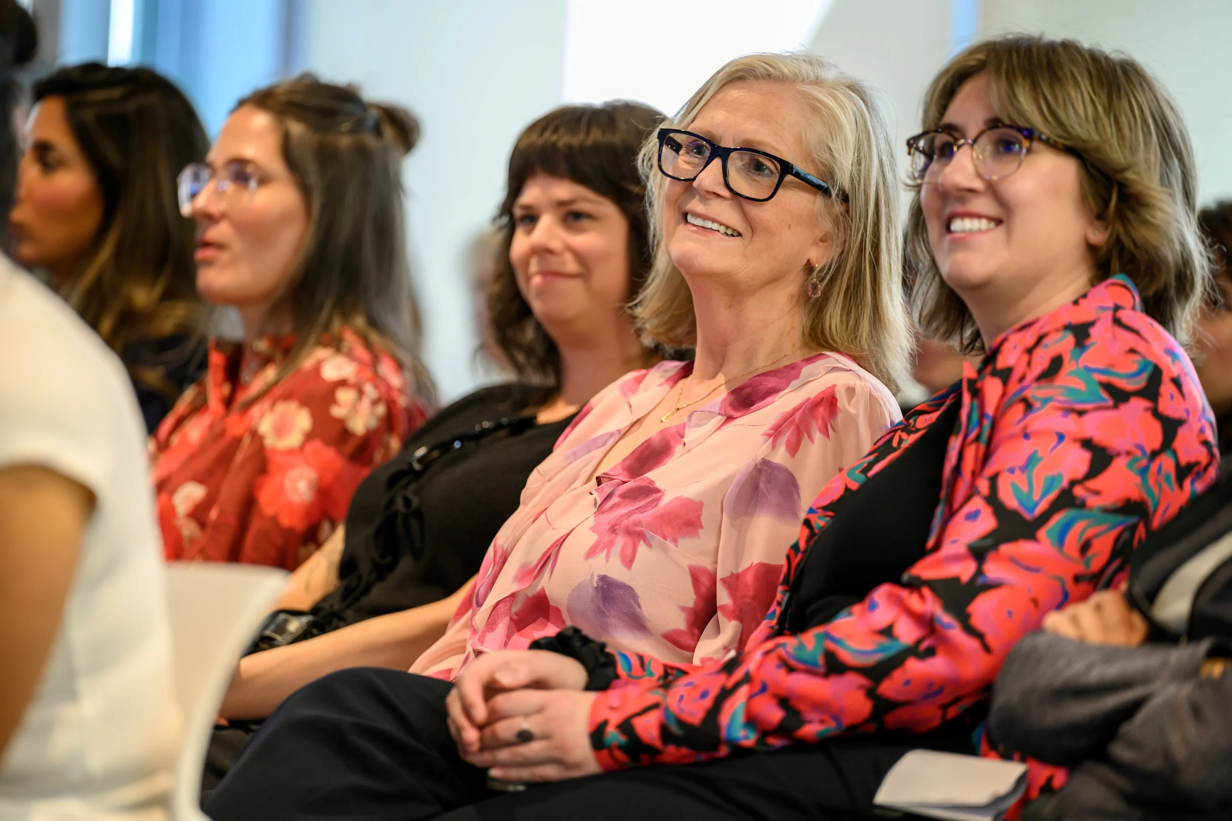 A group of women sitting in a conference or seminar, smiling and listening attentively.