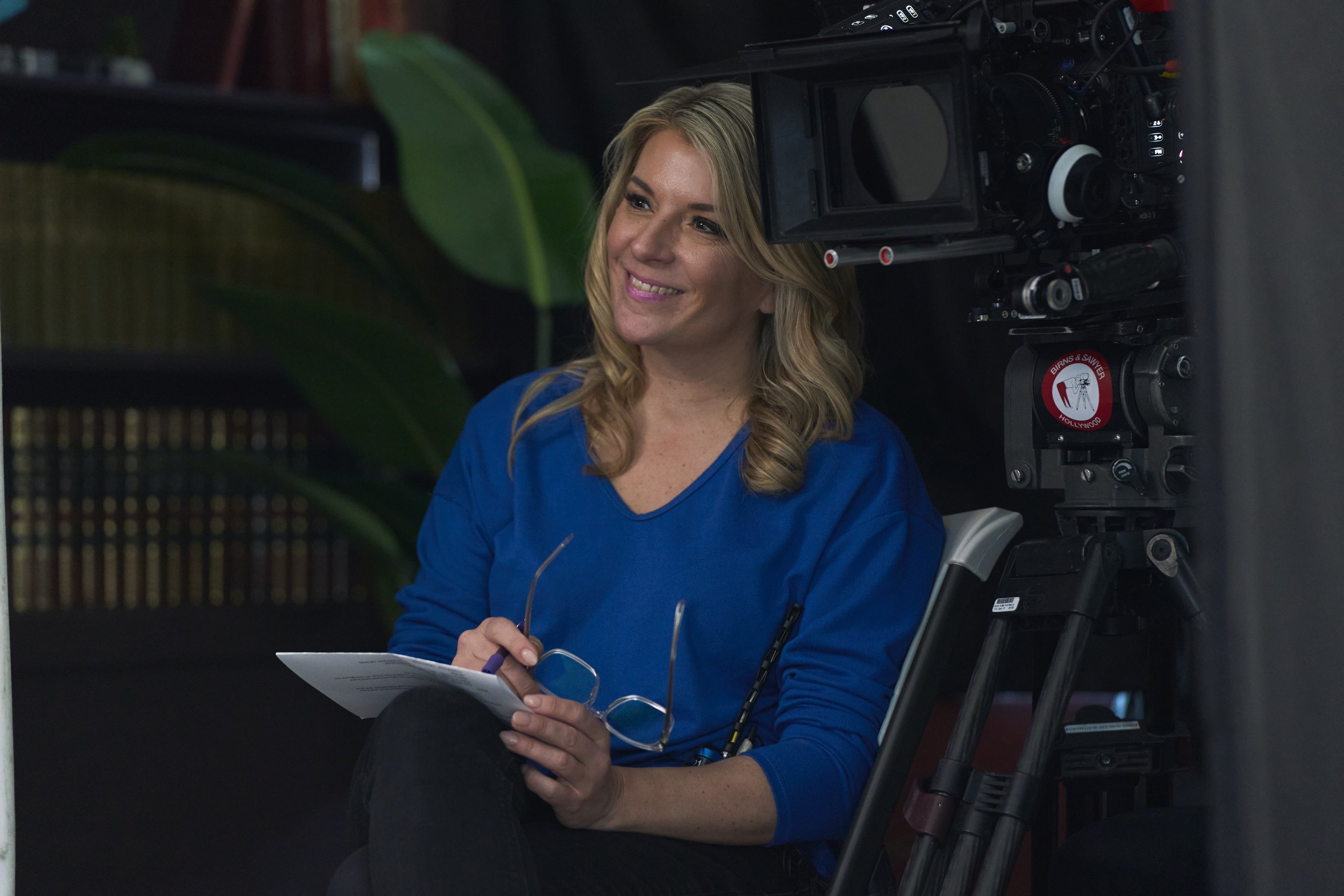 A woman with blonde hair, wearing a blue top, smiling and sitting in front of a film camera, holding glasses and notes.