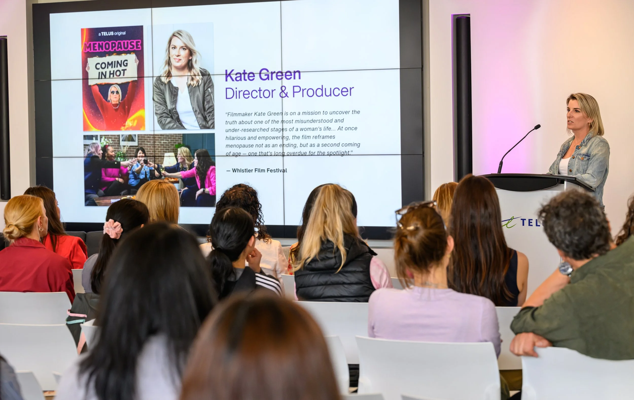 A woman with blonde hair, wearing a denim jacket, speaks at a podium during a presentation in a conference room filled with an audience. A large screen behind her displays her name, Kate Green, and her title as Director & Producer, along with images and text about a film.