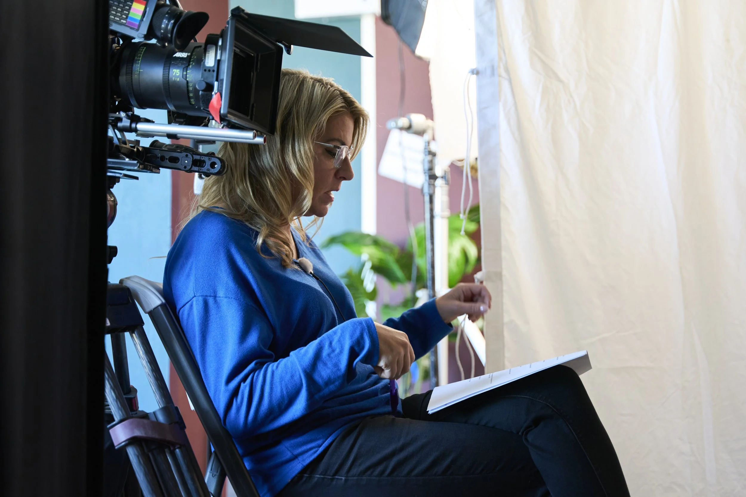 Kate sitting on a chair, working with papers in front of a whiteboard, while a professional film camera is used to record her.