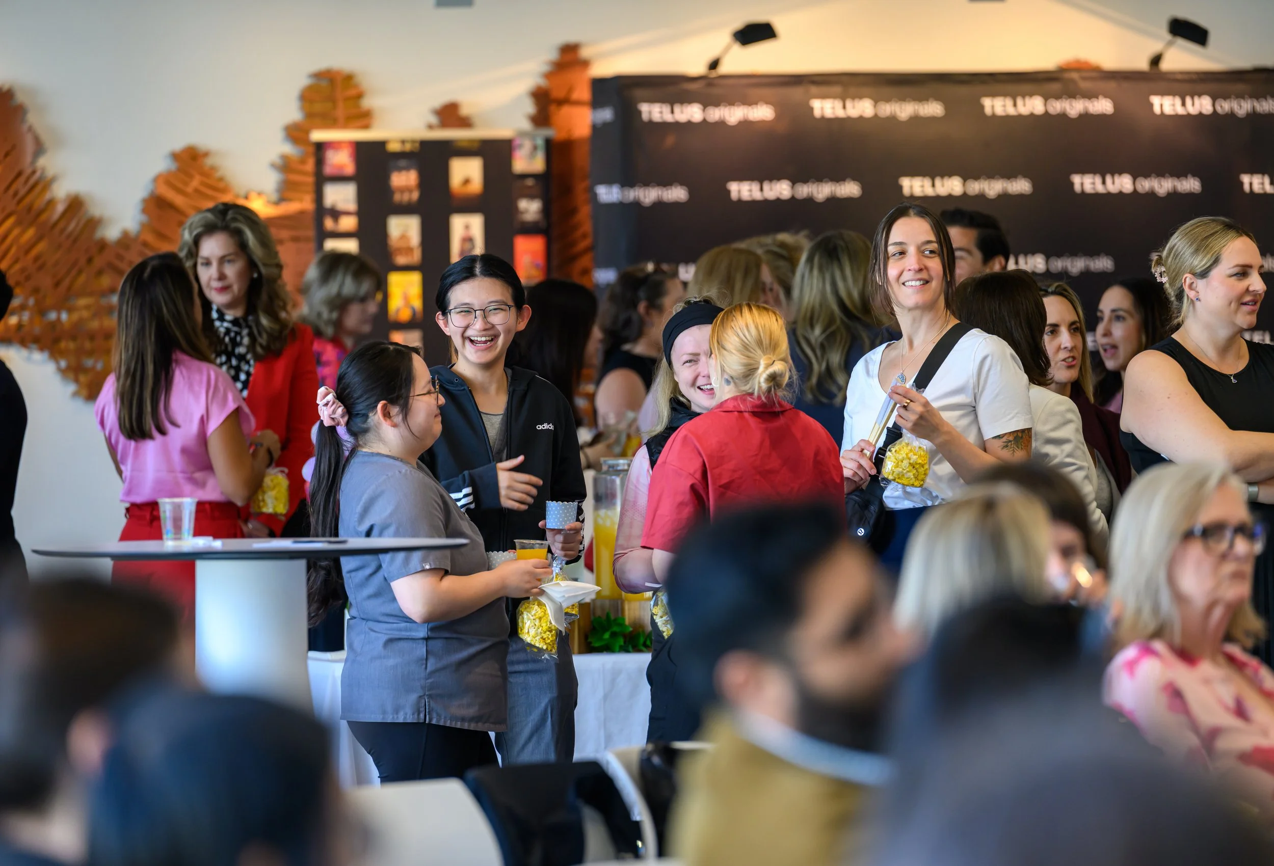 A group of people smiling and chatting at a social event, with some holding popcorn and drinks, in a room with a black banner that reads 'TELUS Originals' and decorative wall art.