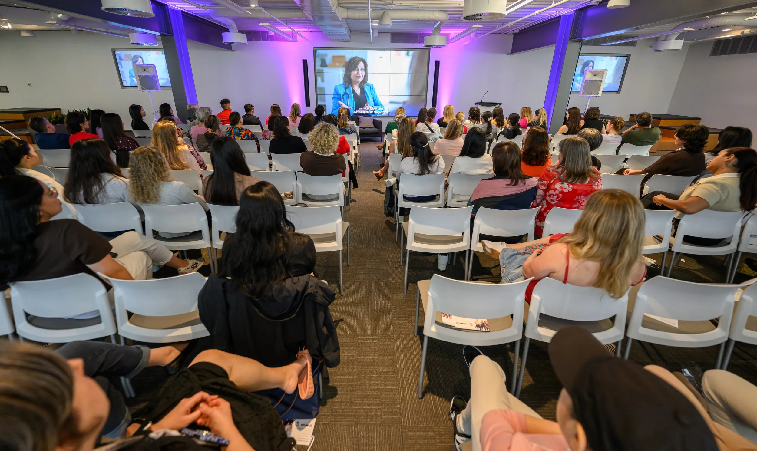 A large group of people attending a presentation or conference in a modern room with purple lighting and a large screen displaying a woman in a blue blazer. Some are seated on white chairs, facing the screen.
