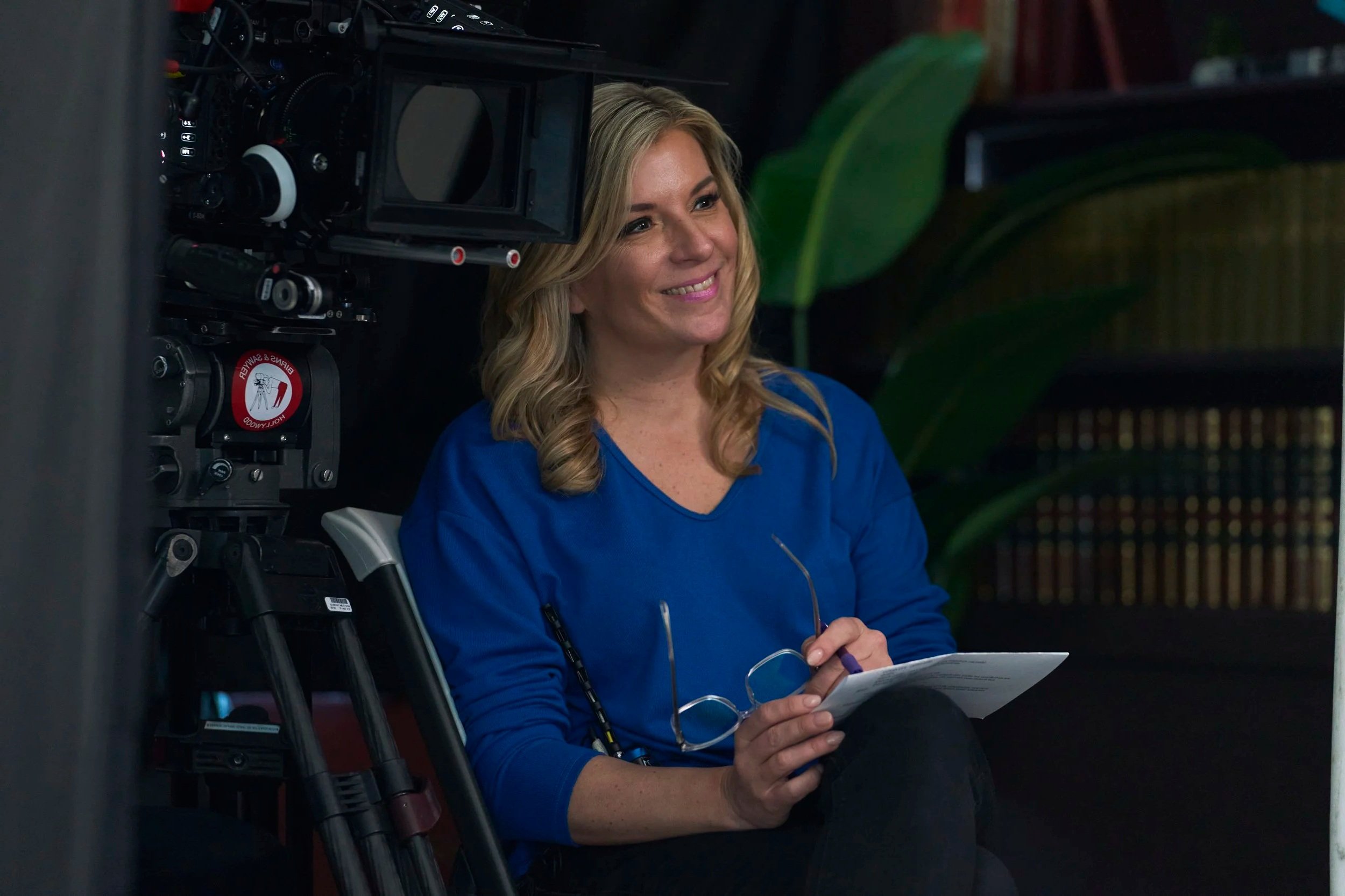 A woman with blonde hair smiling, sitting next to a camera on a tripod, holding a notebook and pen, with large green leaves in the background.