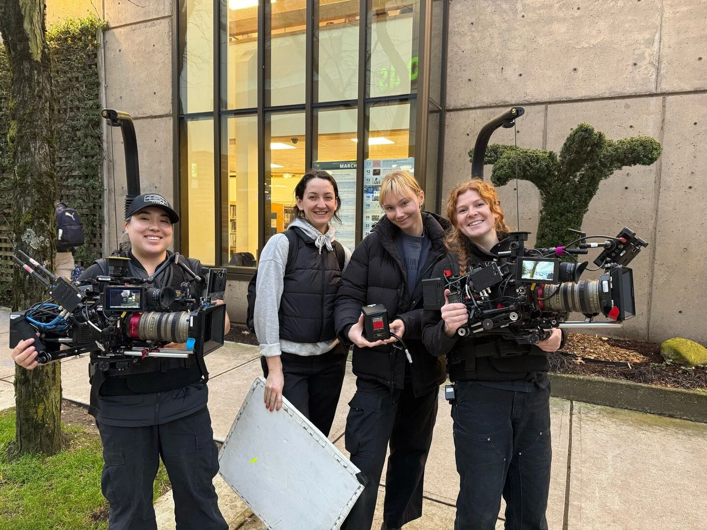 Four women filmmaker crew members standing outdoors in front of a building with large glass windows, holding professional camera equipment and smiling at the camera.