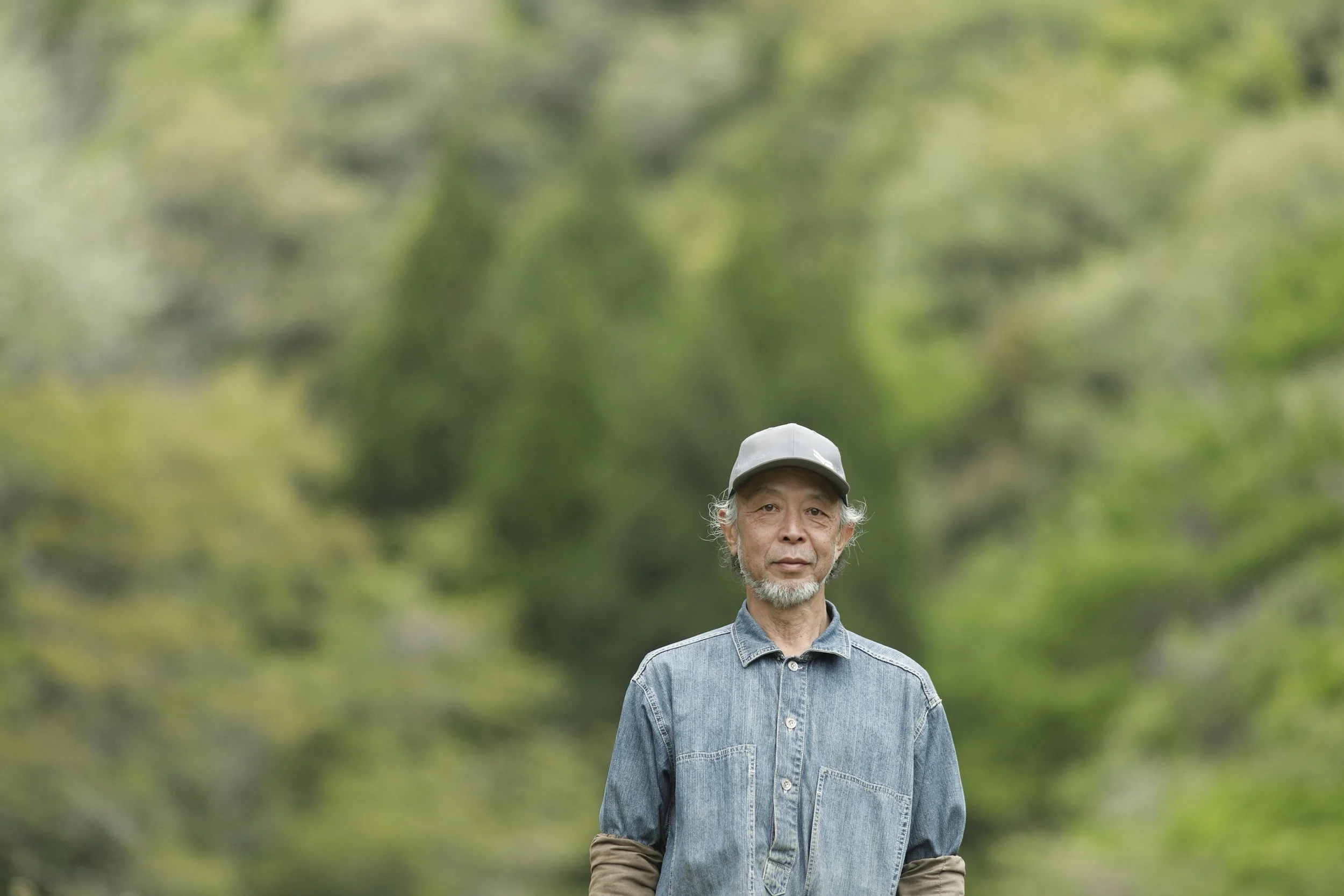 緑の背景の中で、白い帽子をかぶった中年の男性が青いジーンズのシャツを着ている。
