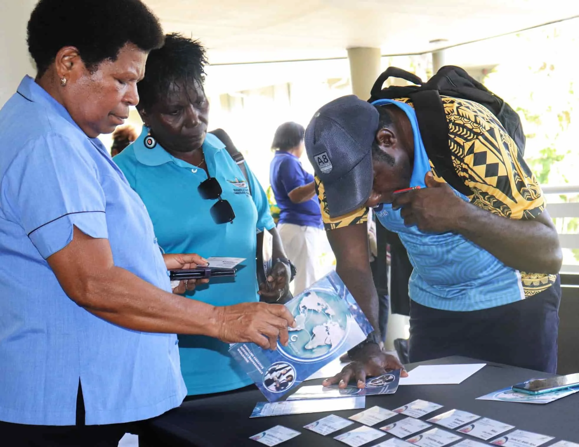 Three people standing around a table with brochures and business cards, one of them signing a brochure, under an outdoor shelter.