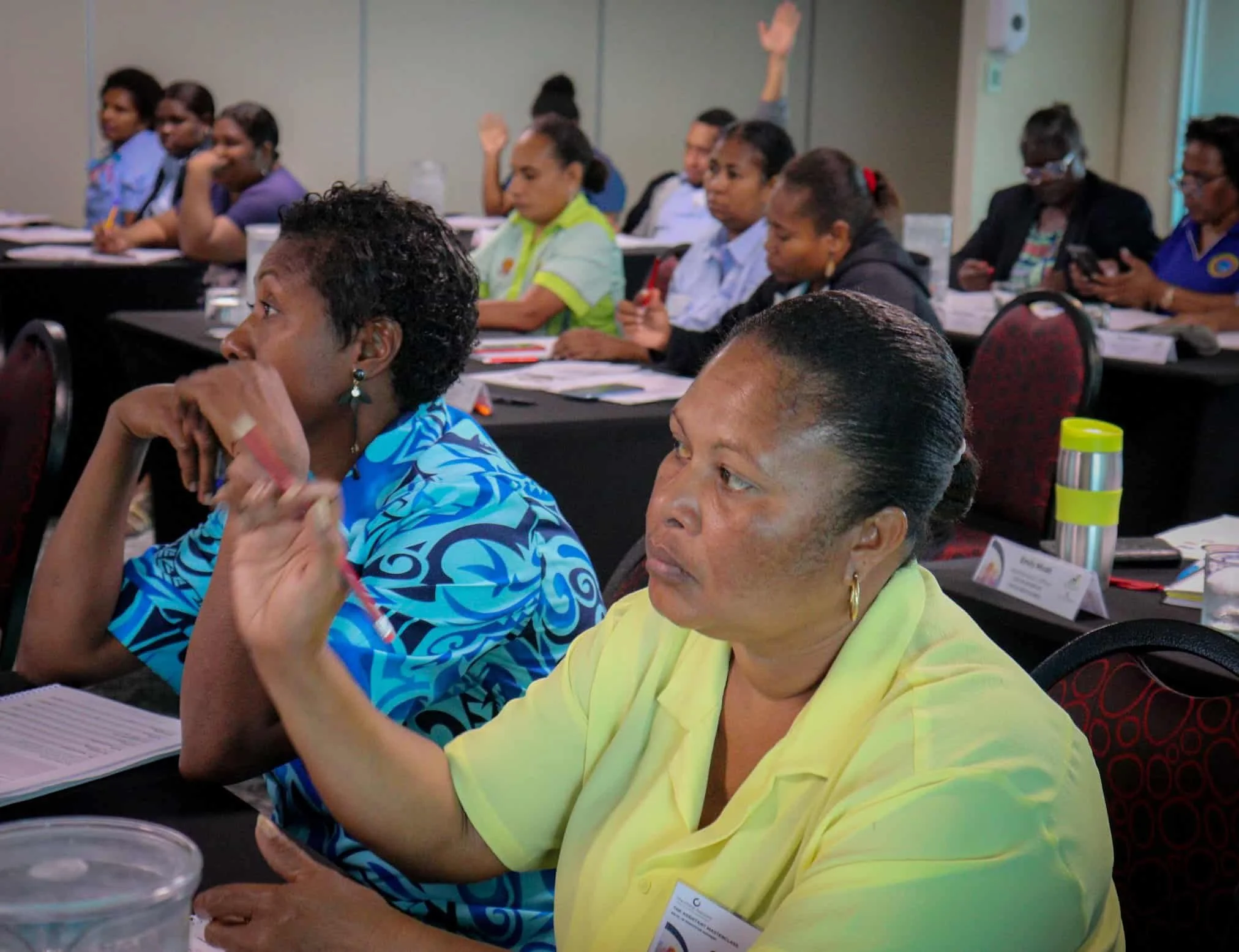 A group of women attending a conference or seminar, sitting at tables with notebooks and drinks, some raising their hands and others listening attentively.
