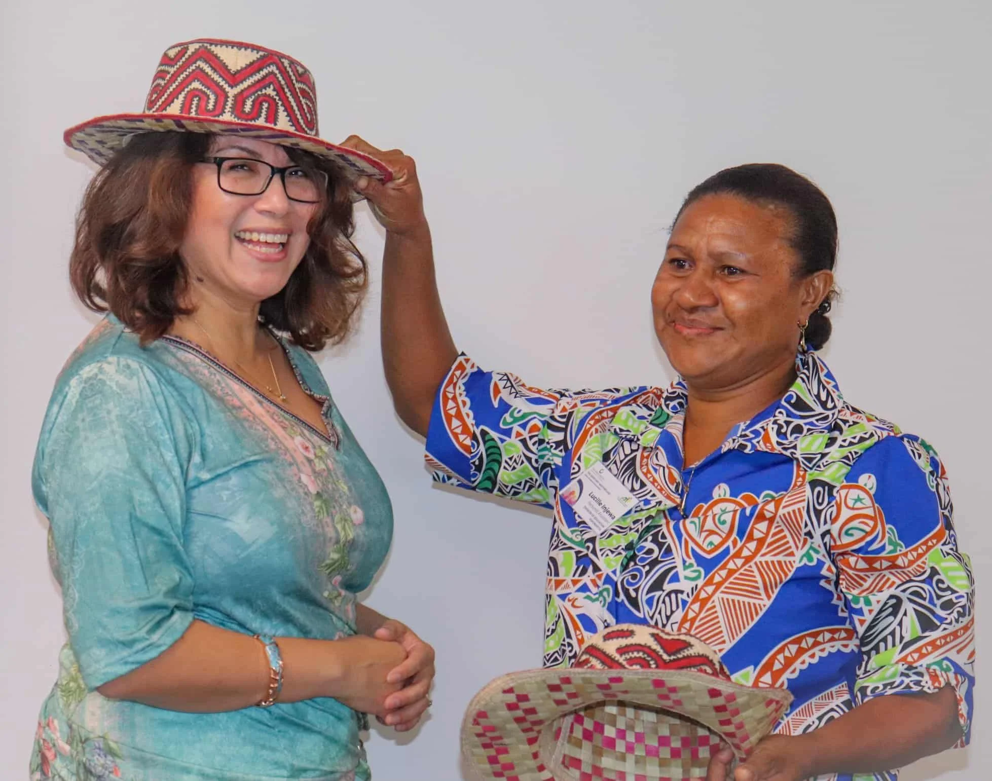 A woman in a colorful embroidered shirt holds a woven hat while another woman in a patterned dress places a wide-brimmed, embroidered hat on her head, both smiling.