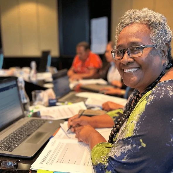 Smiling woman with short curly gray hair and glasses sitting at a conference table with documents and a laptop, other people in the background.