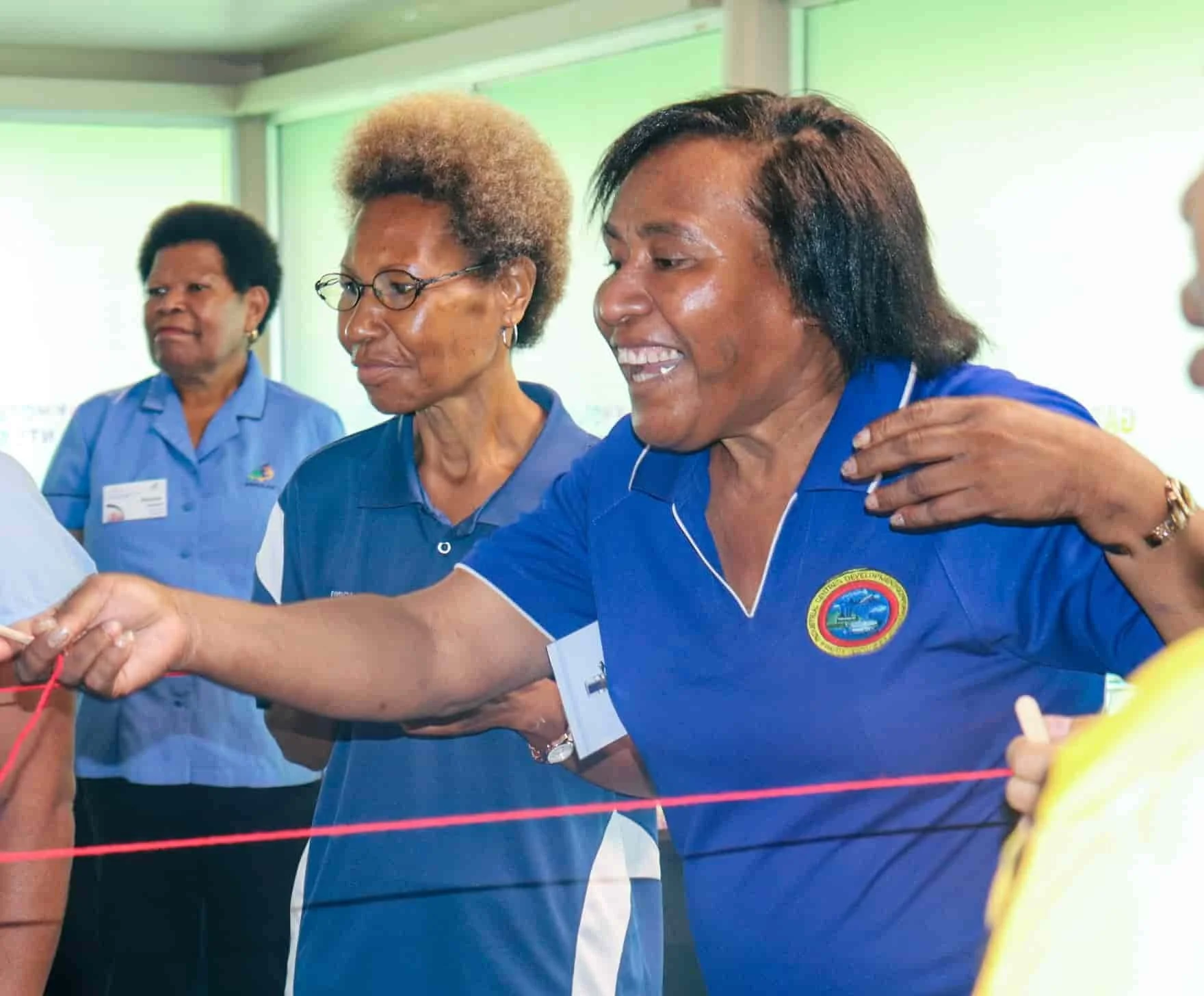Group of women in blue shirts, one smiling woman in the foreground holding a red string during an event or meeting.