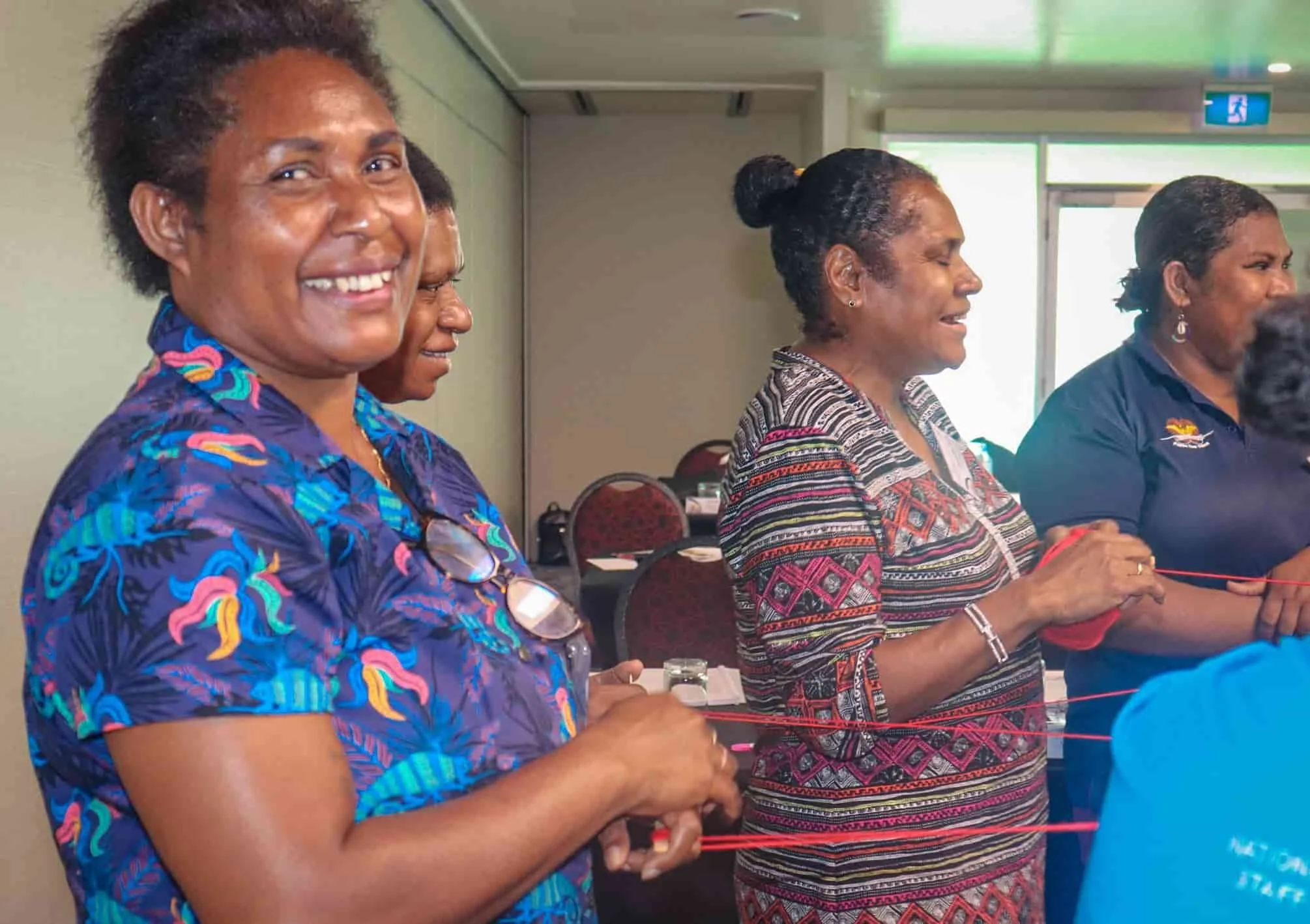 Group of women standing in a conference room, holding a red string, smiling and engaged in an activity.