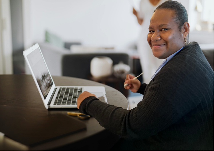 A smiling woman with dark skin and a bun hairstyle, wearing a black blazer, sitting at a desk with an open laptop, in a bright office space.