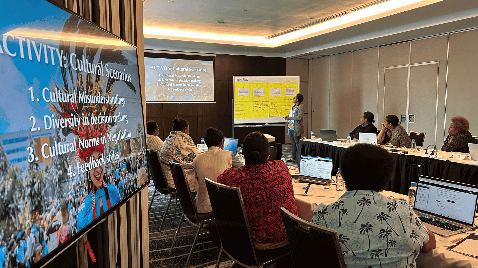A conference room with women seated at a U-shaped table, attending a presentation on cultural scenarios. The presenter stands near a yellow-whiteboard, and a screen displays a slide.