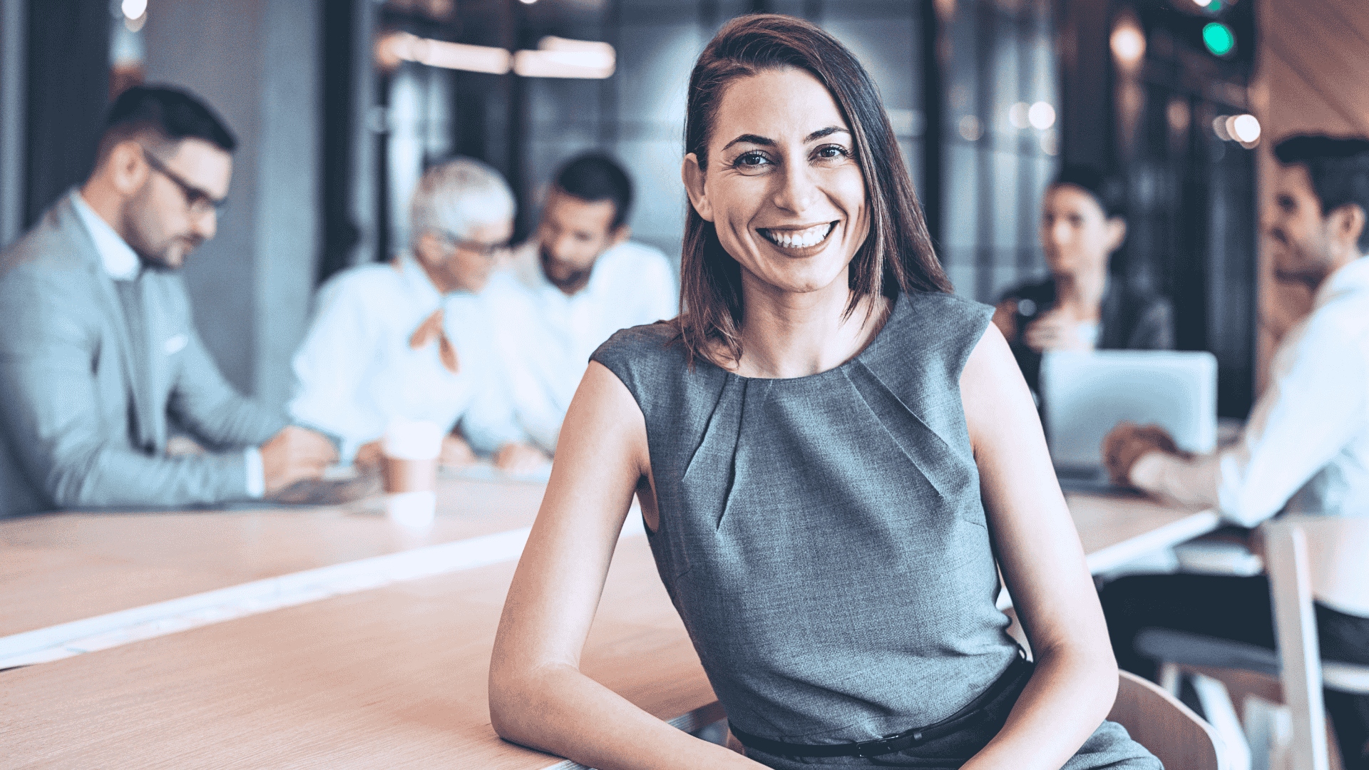 A smiling woman sitting at a conference table in a modern meeting room with five colleagues in the background.