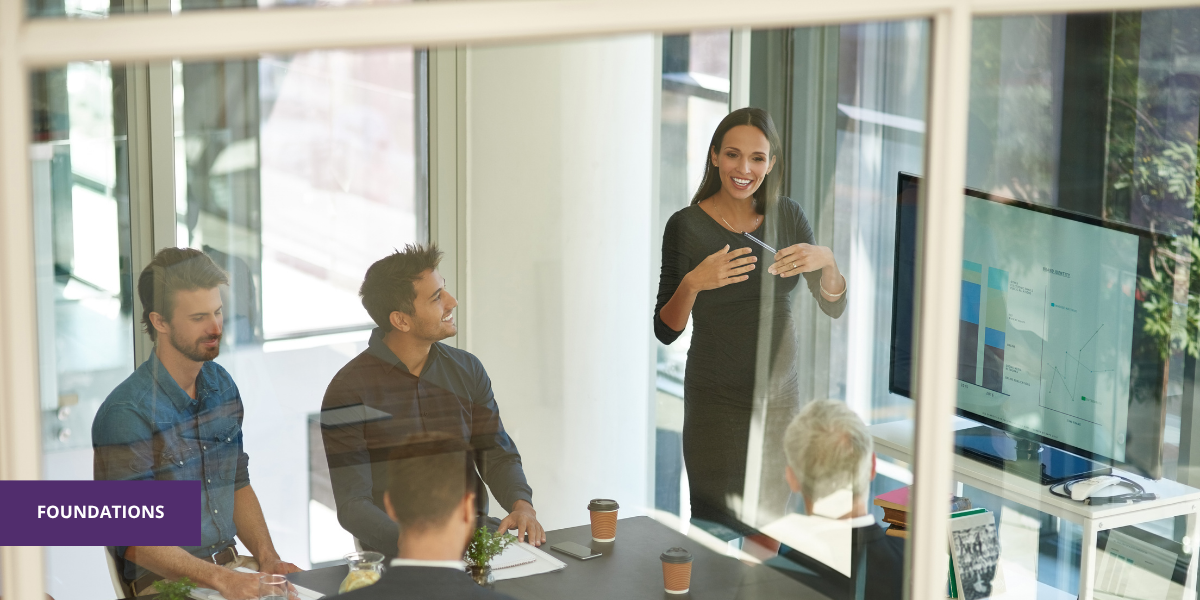 A woman giving a presentation to four colleagues in a glass-walled conference room, with a large screen displaying charts and graphs.