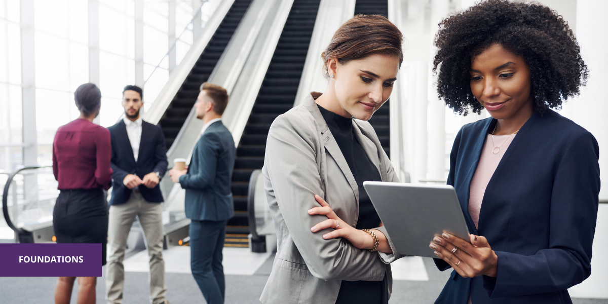 Two women in business attire looking at a tablet, with a group of three people talking in the background near an escalator in a modern office building.