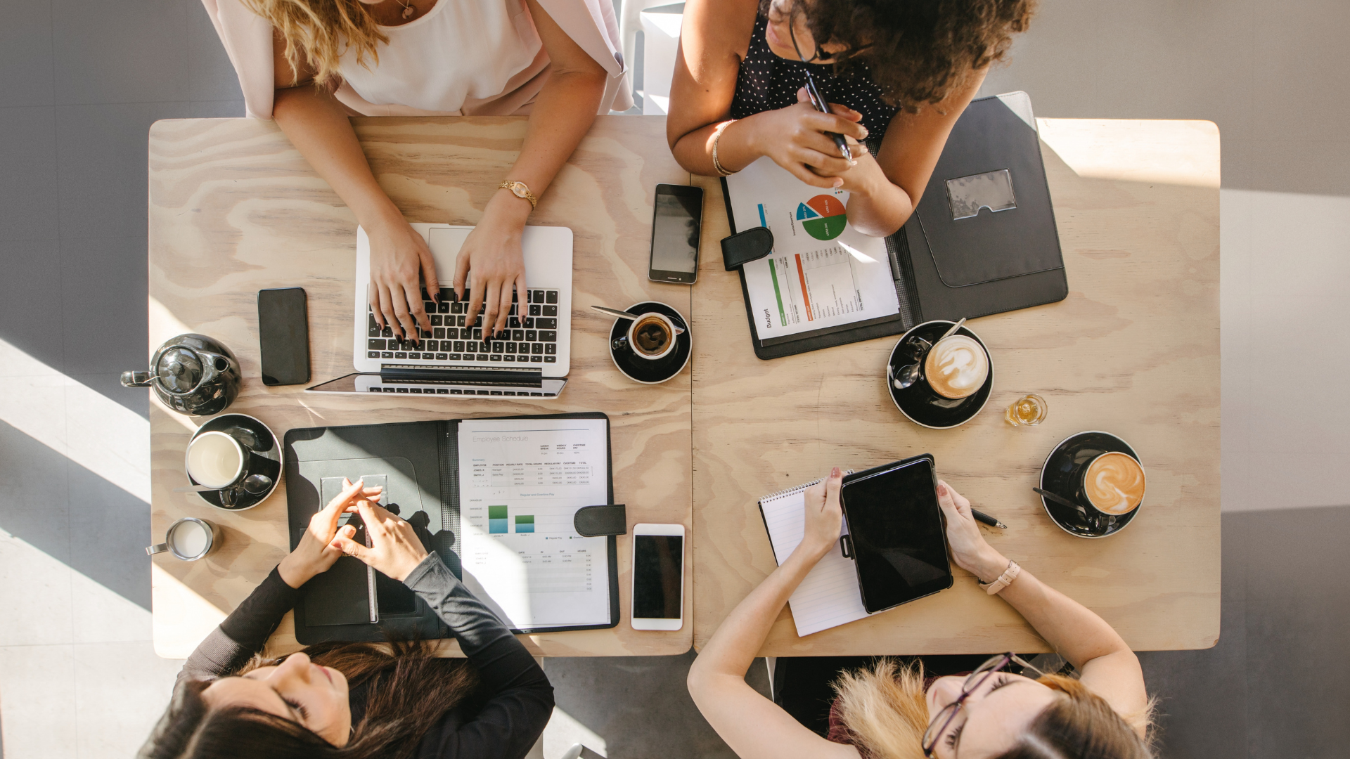 Overhead view of a group of people working at a wooden table with laptops, tablets, smartphones, notebooks, coffee cups, and glasses of water.