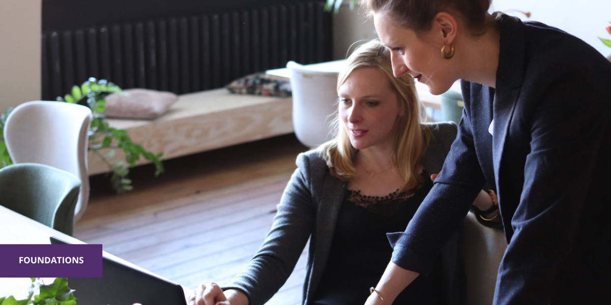 Two women in a meeting room looking at a laptop, one is seated and the other is standing and leaning over to view the laptop screen, both smiling.
