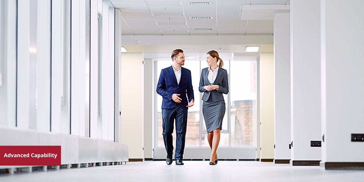 A man and woman in business attire walking and talking in an office hallway.