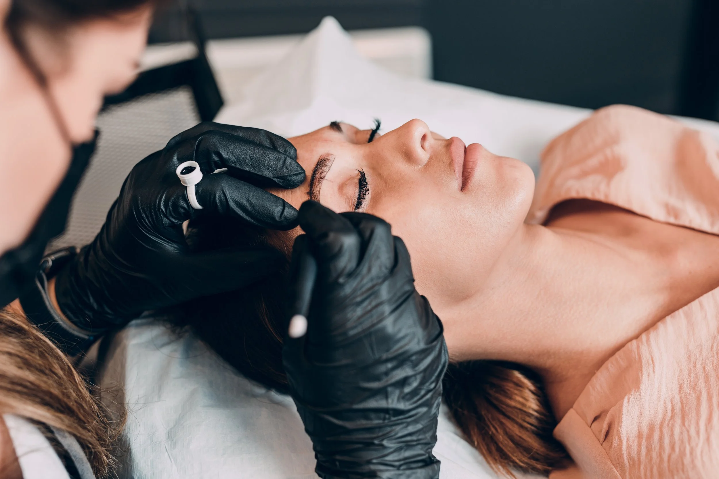 A woman receiving a facial treatment with closed eyes, lying on a bed, while a skincare professional in black gloves applies a product or mask to her forehead.