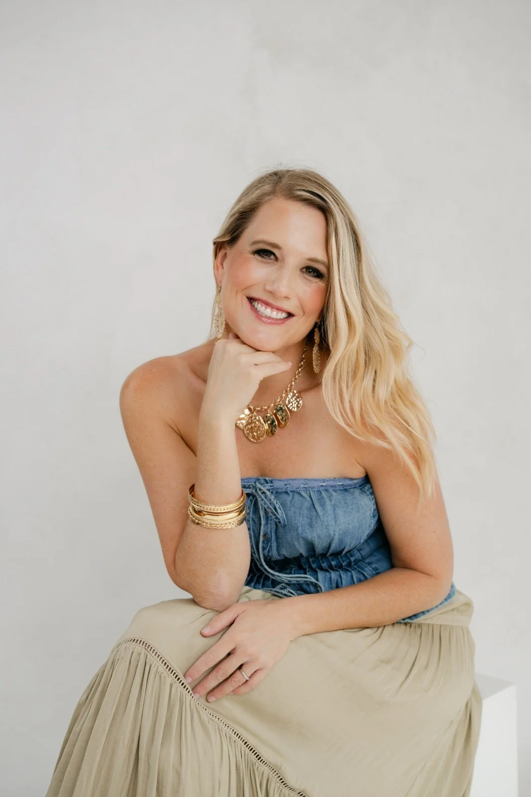 A smiling woman with blonde hair, wearing gold jewelry, a strapless blue top, and a beige skirt, sitting against a white background.