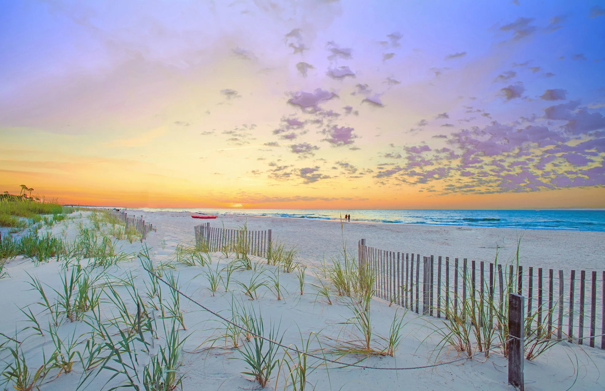 A scenic beach at sunset with the sky painted in soft pastel colors. Sea oats and a wooden fence line the sandy path leading to the shoreline. Two people are walking along the water's edge, and a red boat is resting on the sand near the water.