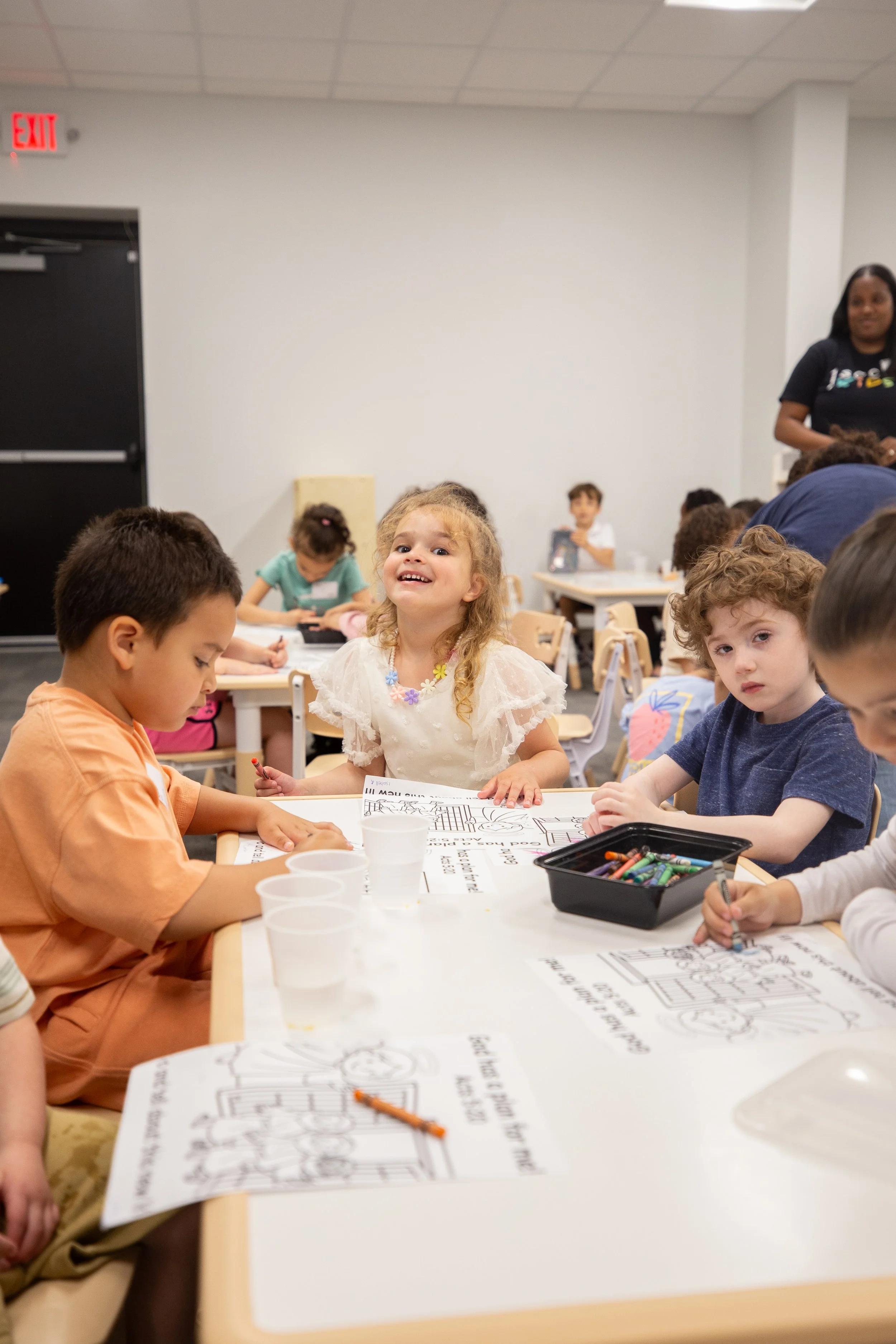 Children participating in a coloring activity in a classroom, sitting at tables with coloring sheets and crayons, some smiling and engaged.