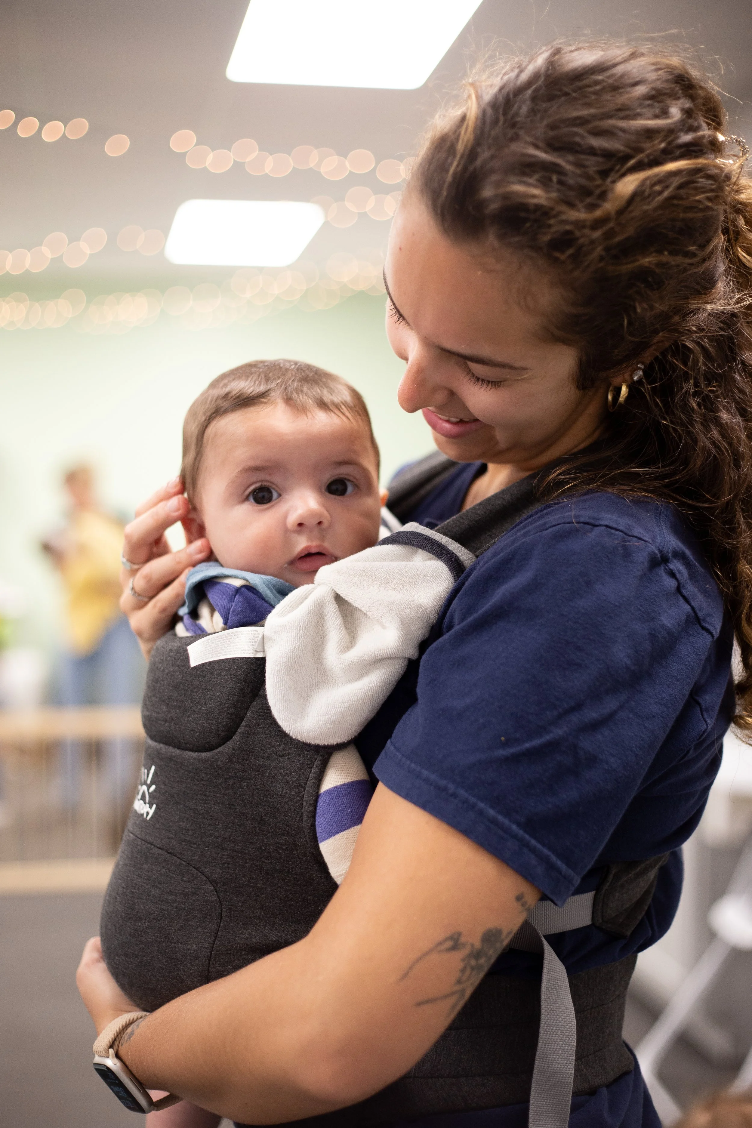 A woman with curly brown hair smiling while holding a baby with large wide eyes and short brown hair, set indoors with blurred lights in the background.