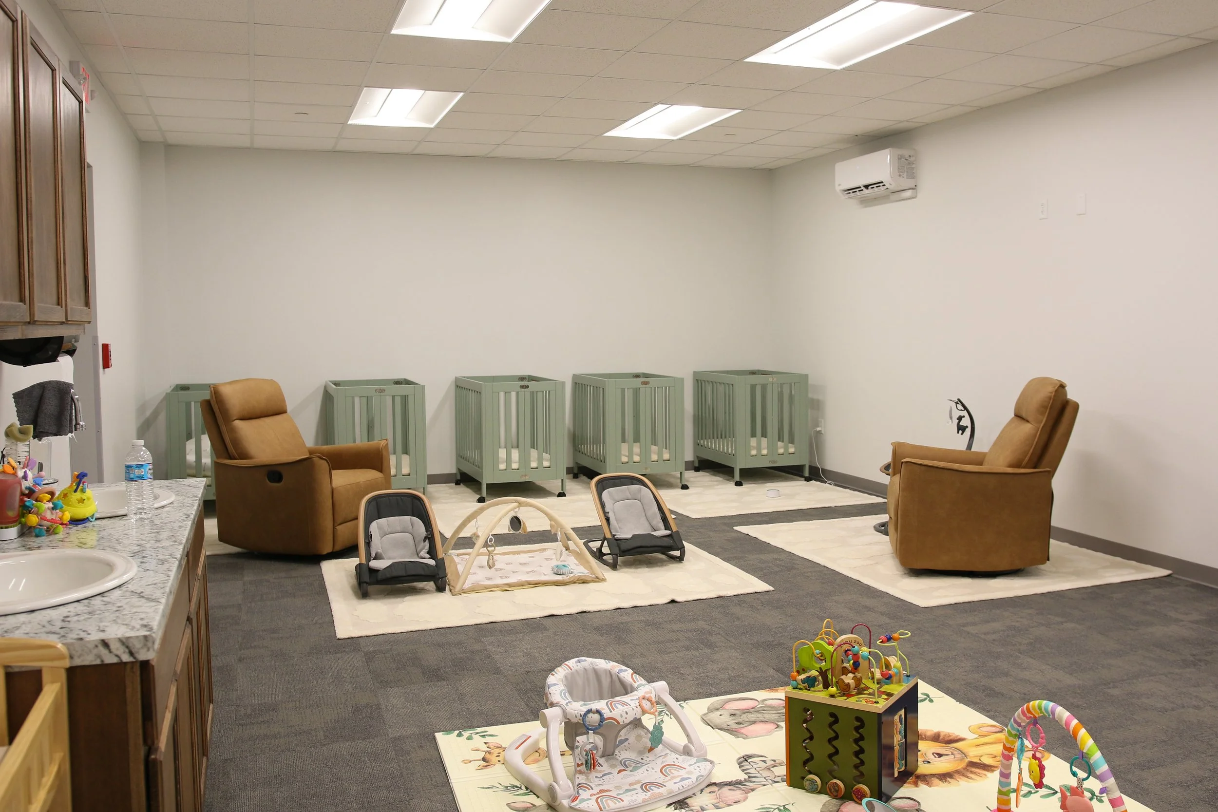 A room with a white ceiling, beige walls, and a gray carpeted floor containing baby furniture, chairs, and toys, indicating a daycare or playroom setting.