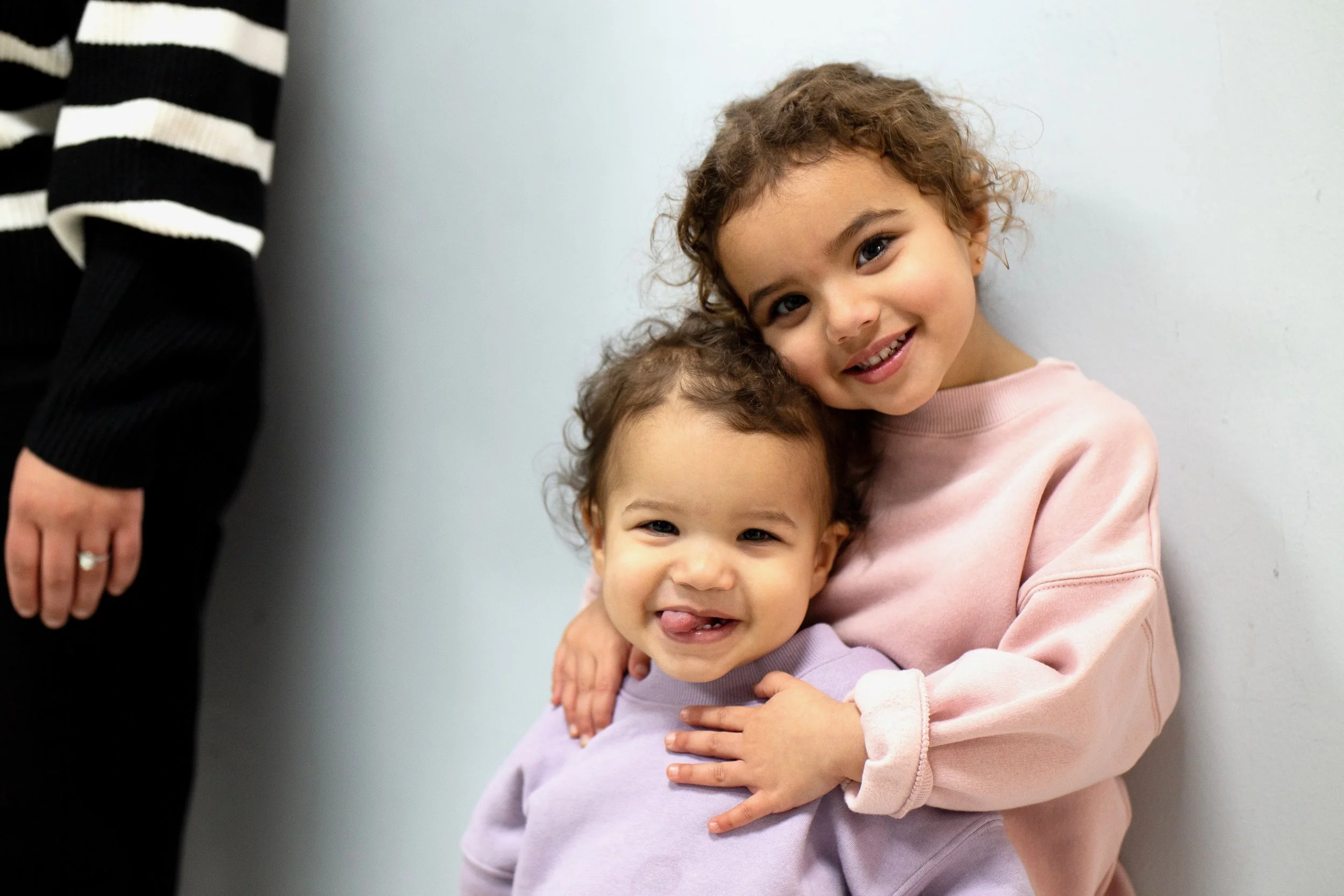 Two young girls hugging and smiling, one in a pink sweatshirt and the other in a light purple sweatshirt, against a plain white background.