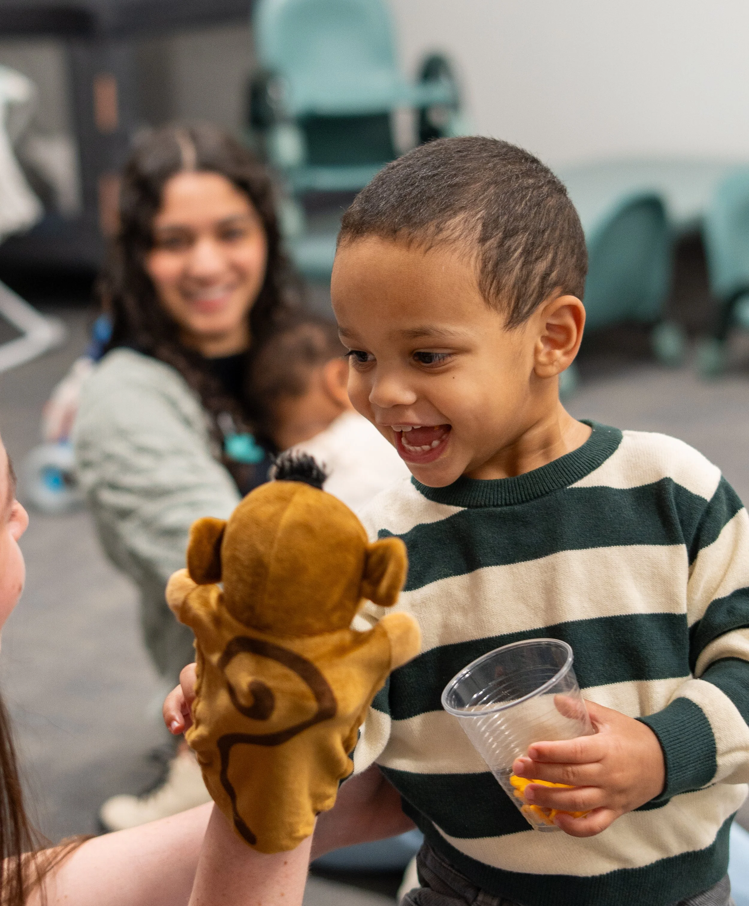 A young boy with a short haircut and a striped sweater smiling and showing his teeth, holding a clear plastic cup filled with yellow snacks, while an adult with curly hair and a smile stands beside him. The person is also holding a large stuffed animal resembling a lion. The background appears to be an indoor setting with chairs and a table.