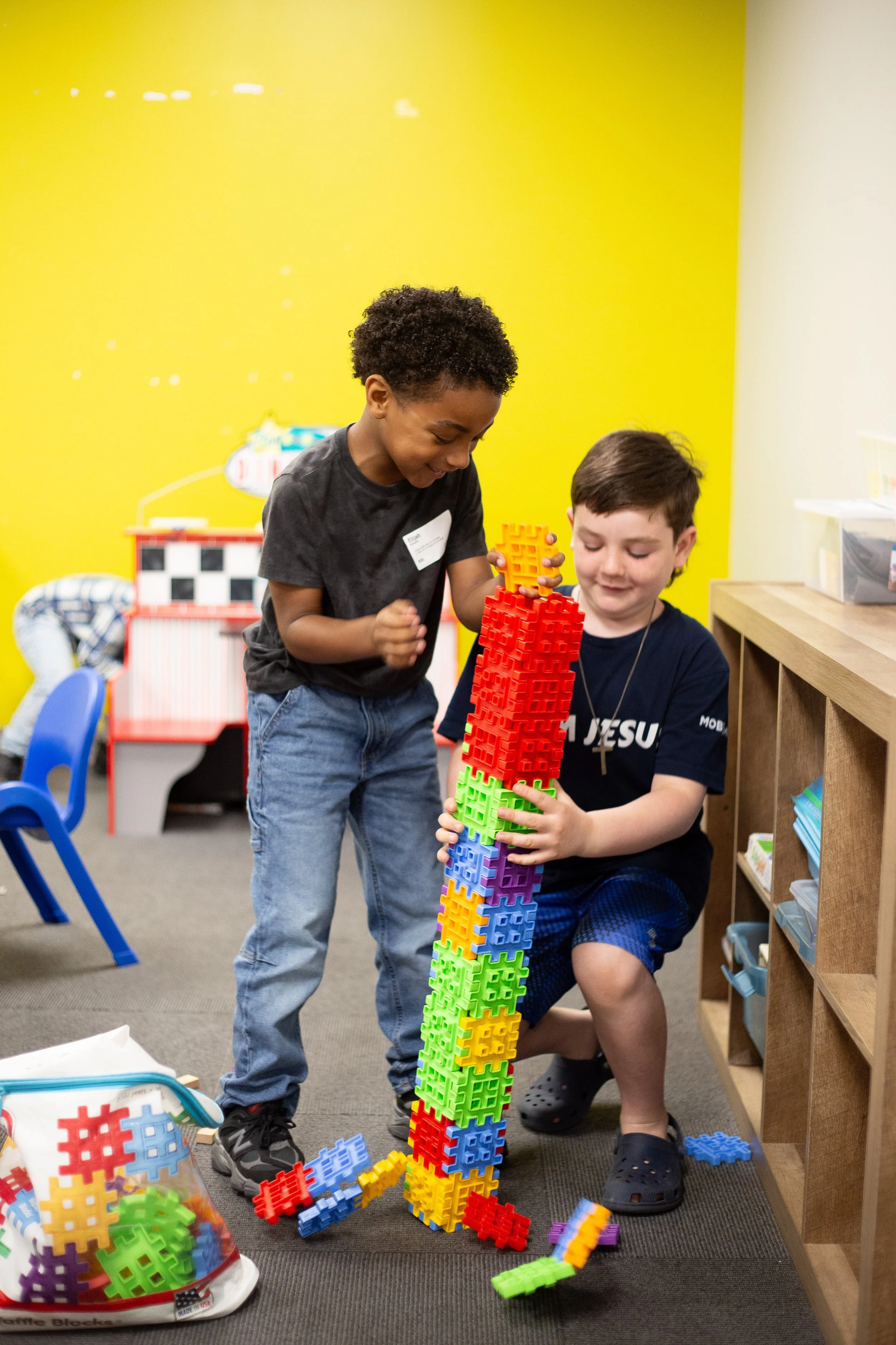 Two boys play with large interlocking building blocks, stacking them high in a room with a yellow wall.